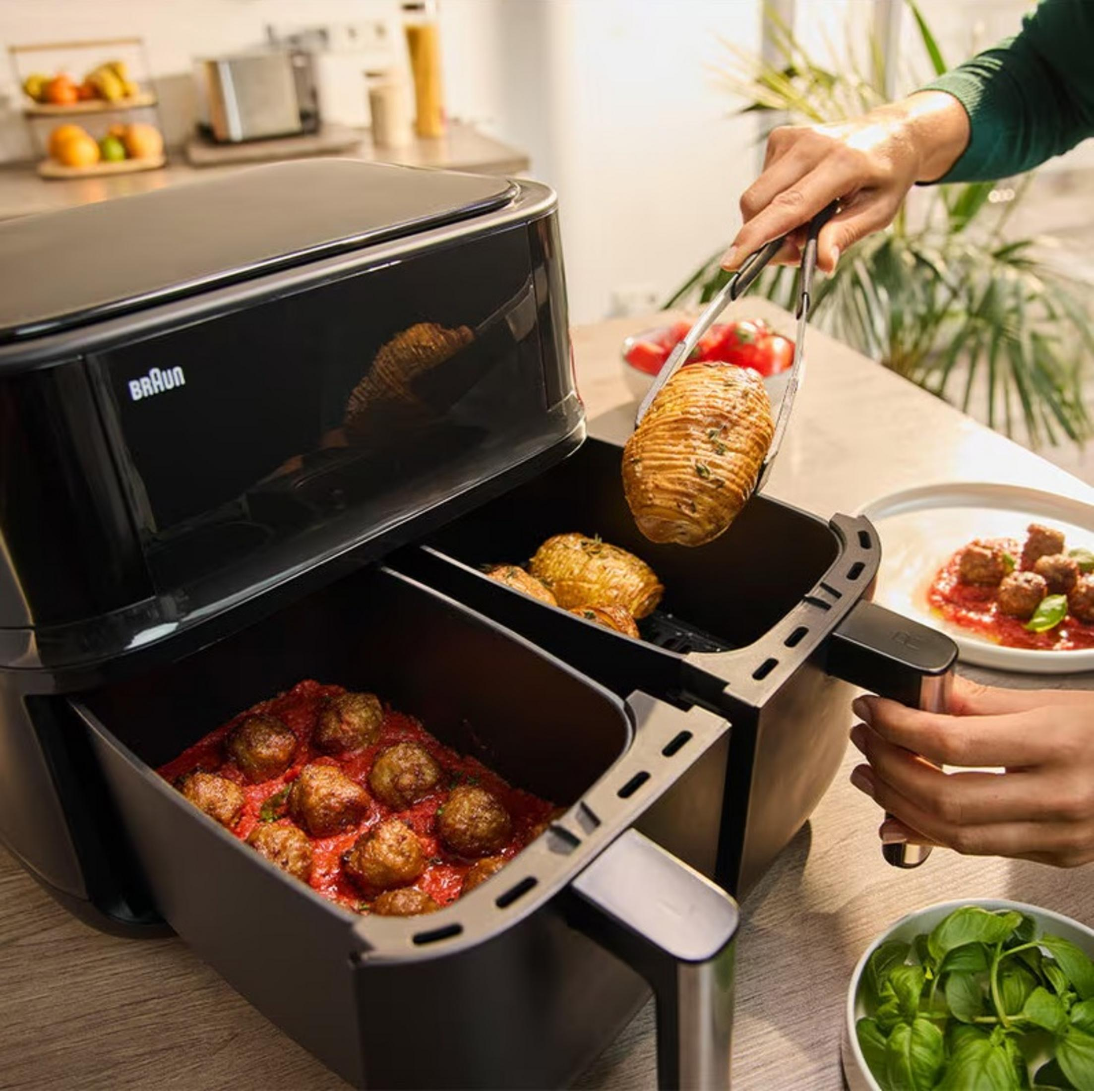 Een airfryer met eten erin, een persoon die een aardappel met een tang eruit haalt. Andere voedingsmiddelen zijn aanwezig.