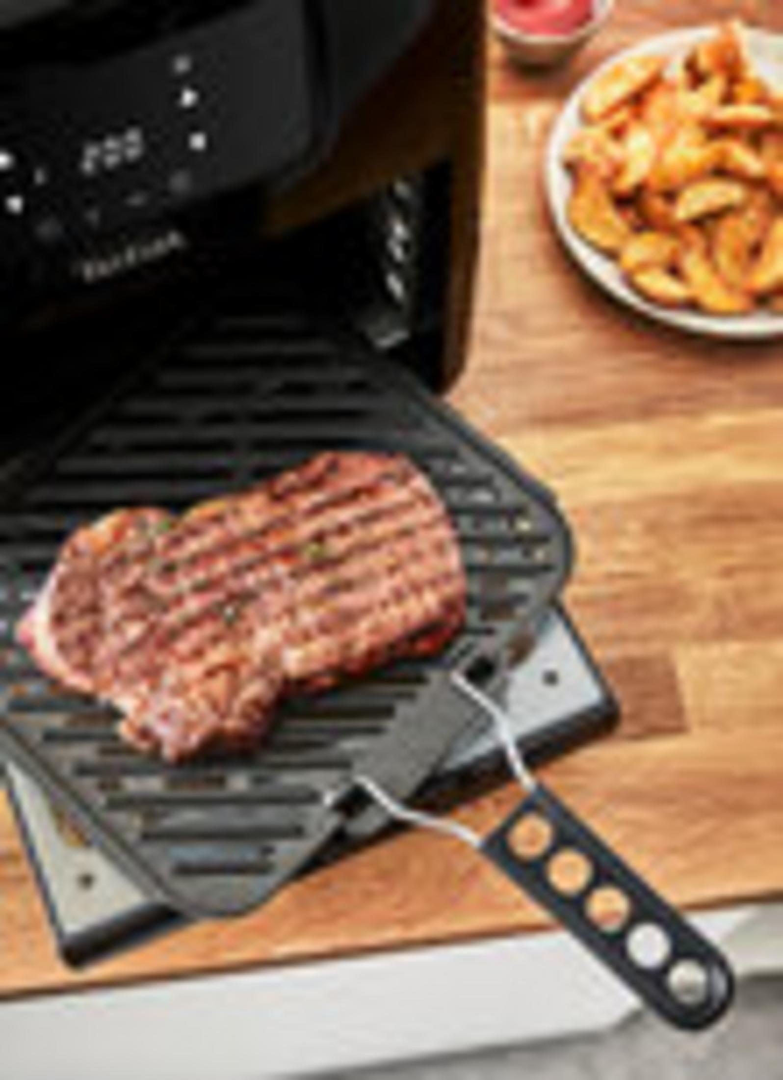 Grillowany stek na patelni grillowej, frytkownica po lewej, frytki i sos w tle, na drewnianym stole.