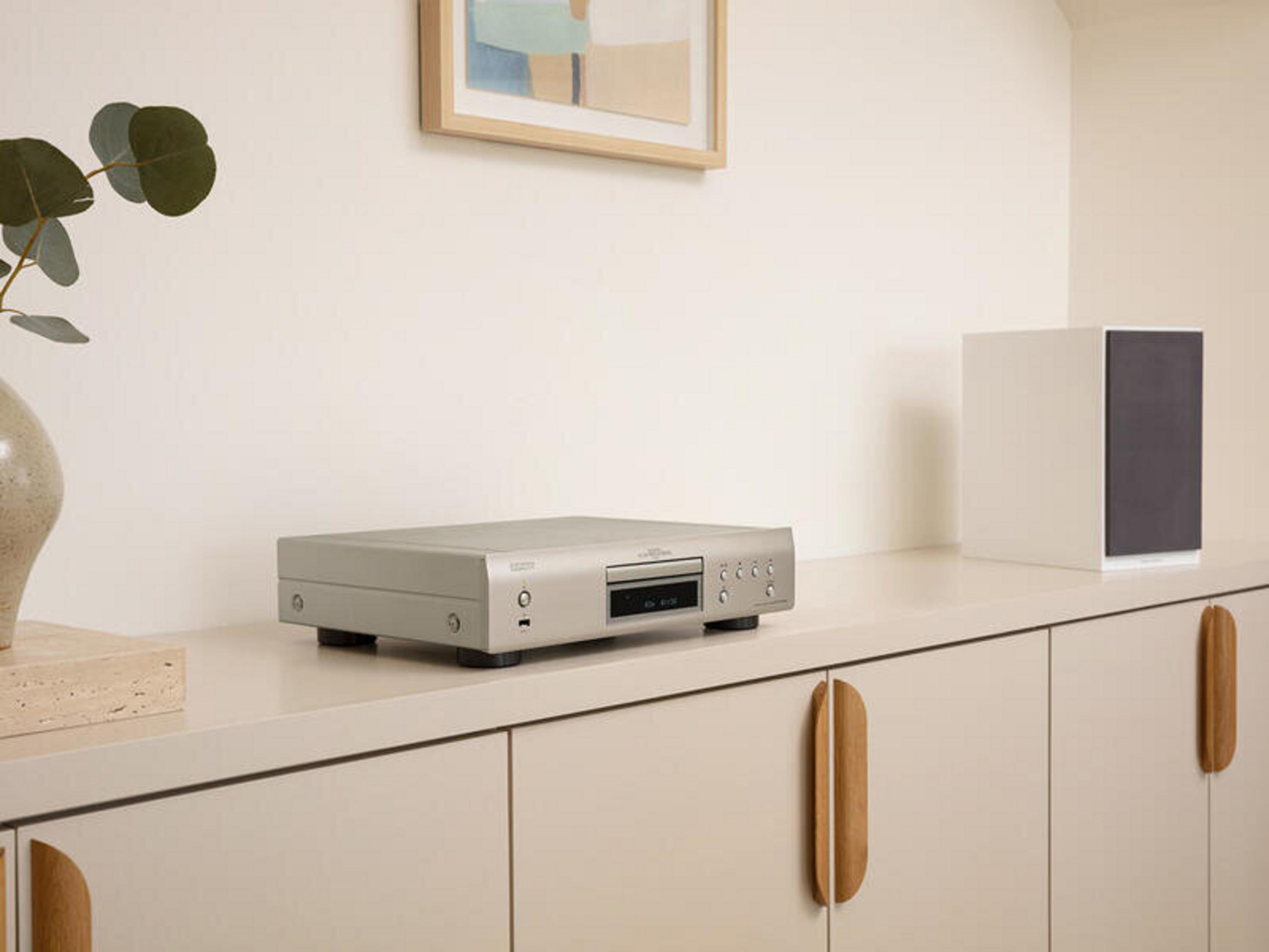 Silberner CD-Player auf weißem Schrank. Ein weißer Lautsprecher und Kunstwerk im Hintergrund.