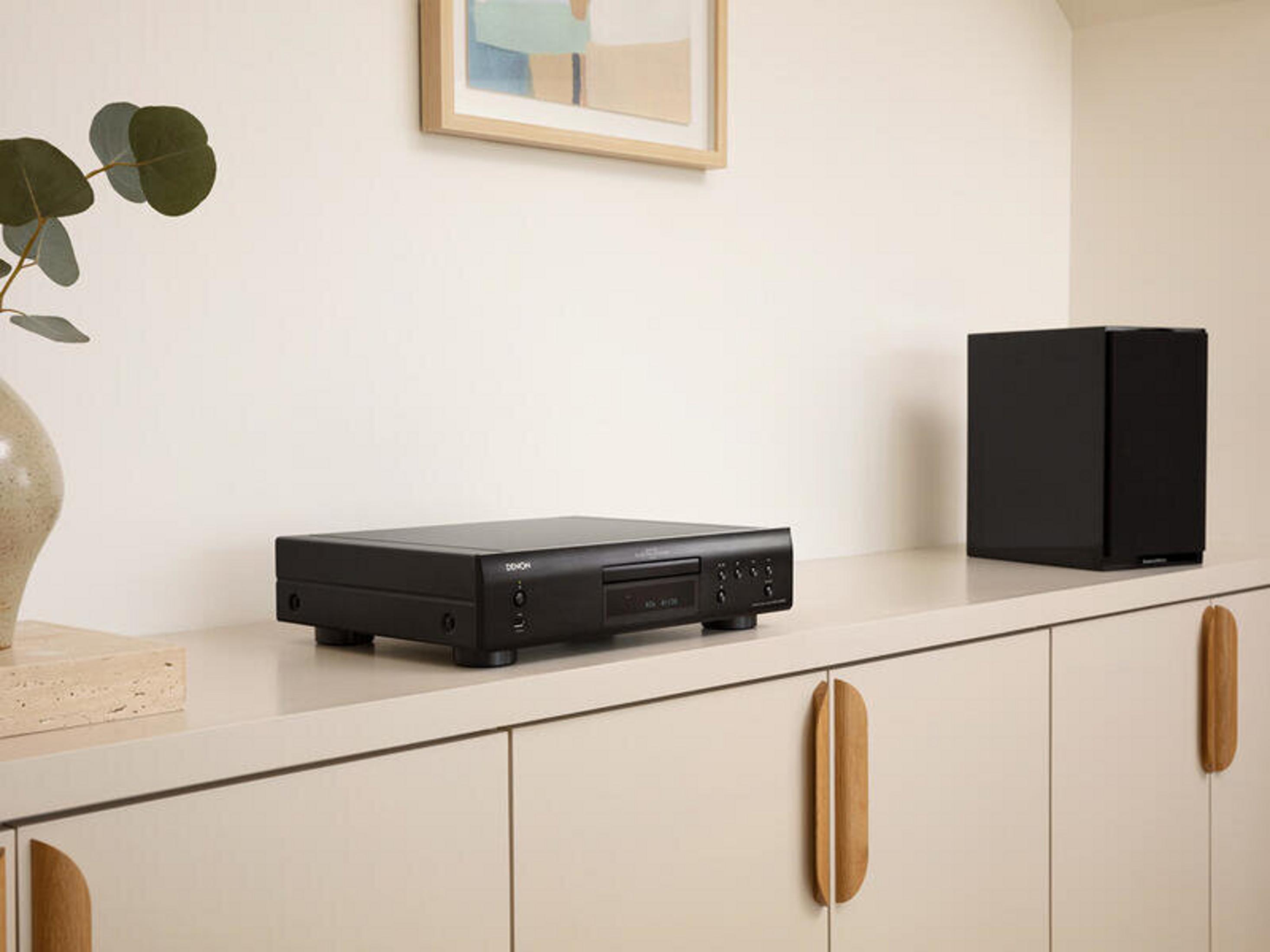 Un reproductor de CD negro, un altavoz negro y un jarrón con hojas están sobre un mueble blanco.