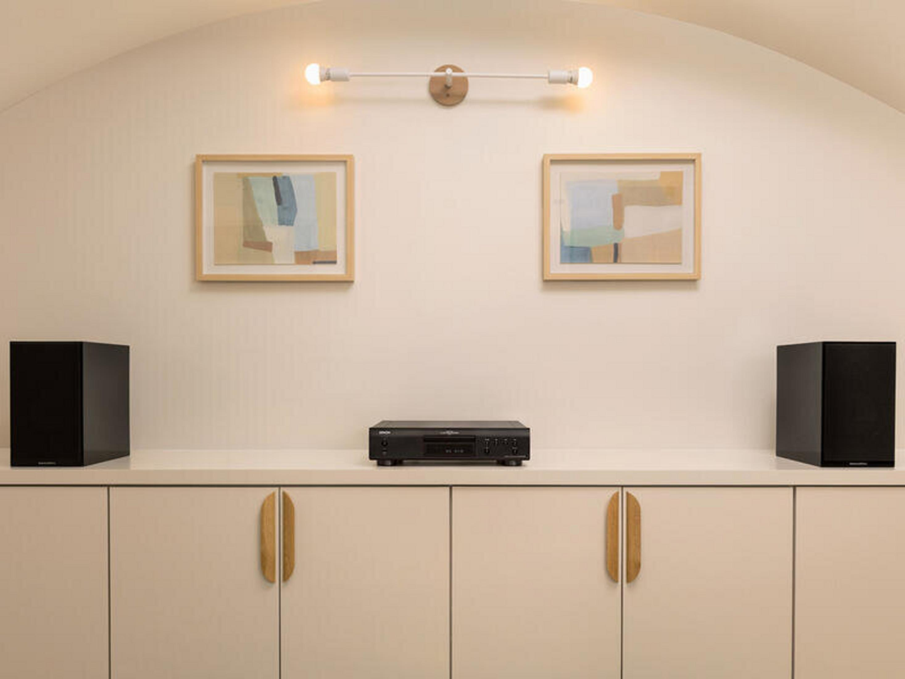 Una habitación con paredes crema, arte, altavoces y un reproductor. Hay lámparas y un armario con tiradores de madera.