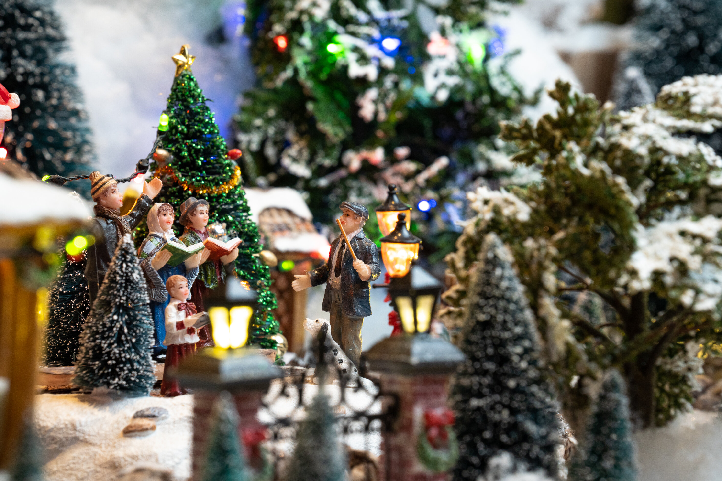 Scène de Noël miniature avec des personnages chantant des chants, un sapin et des arbres et un sol enneigés.