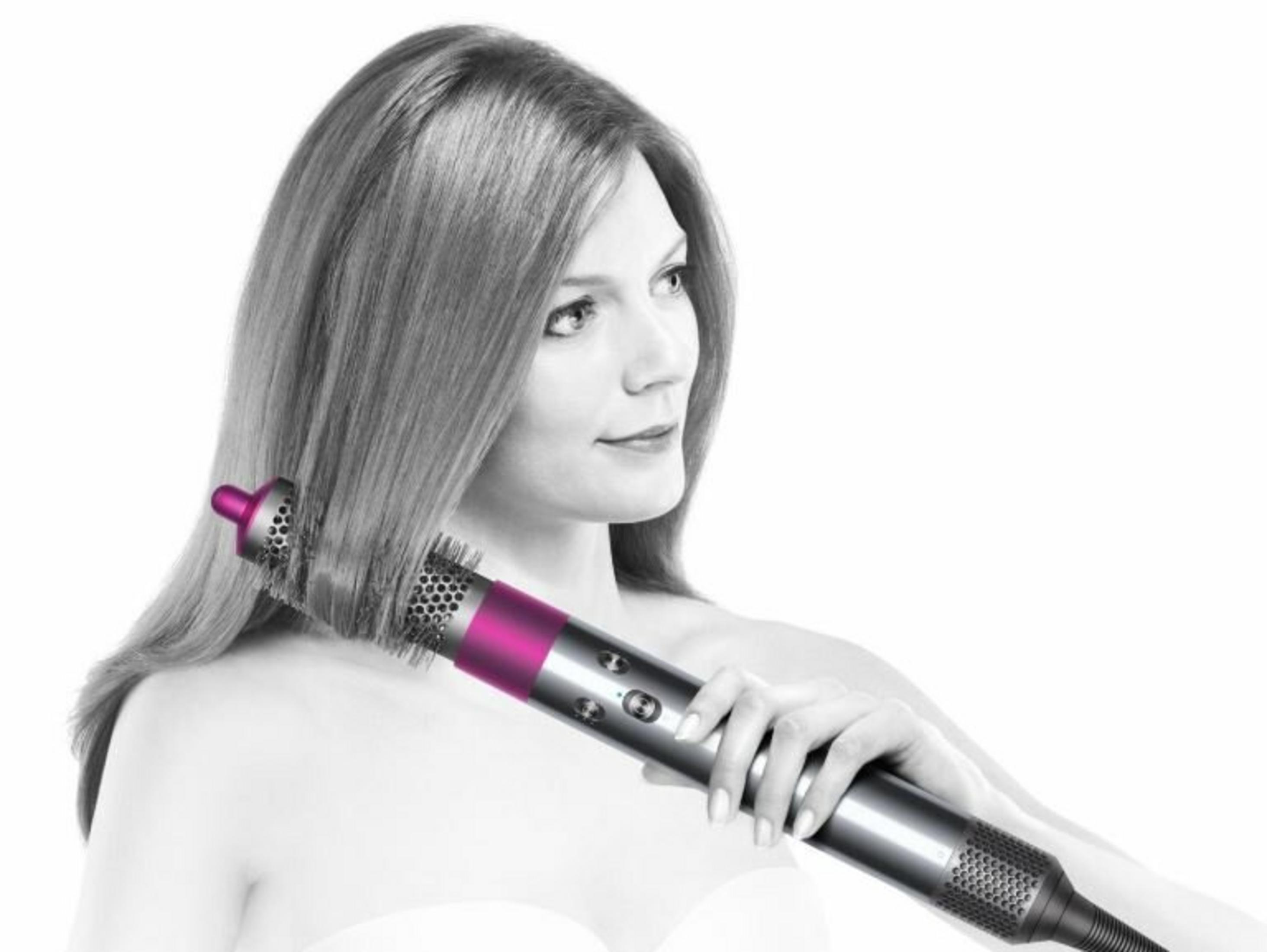 Eine Frau benutzt ein silbernes und pinkfarbenes Haarstyling-Werkzeug. Sie hat langes Haar. Der Hintergrund ist weiß.