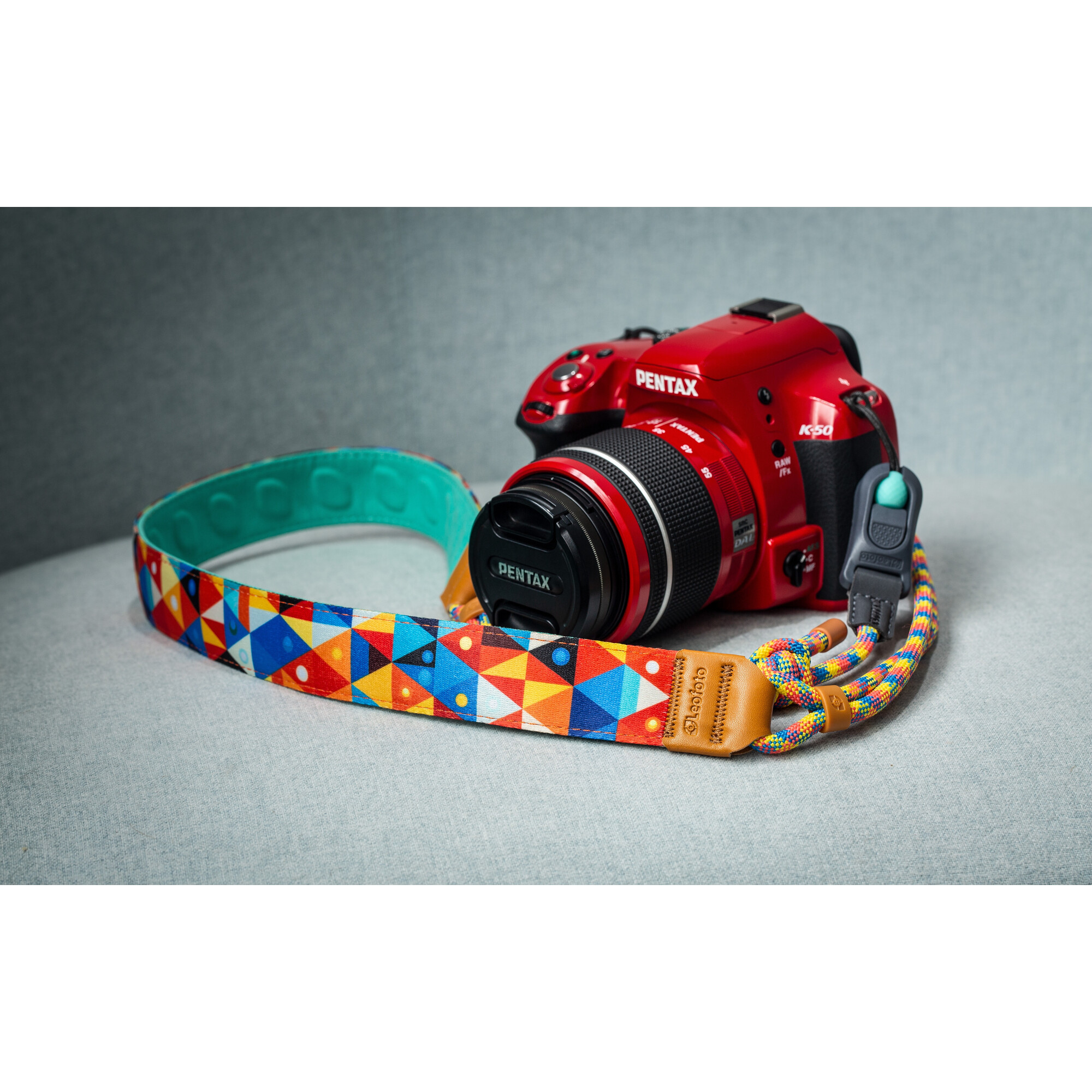Eine rote Pentax-Kamera mit schwarzem Objektiv und farbenfrohem Gurt liegt auf grauer Oberfläche.