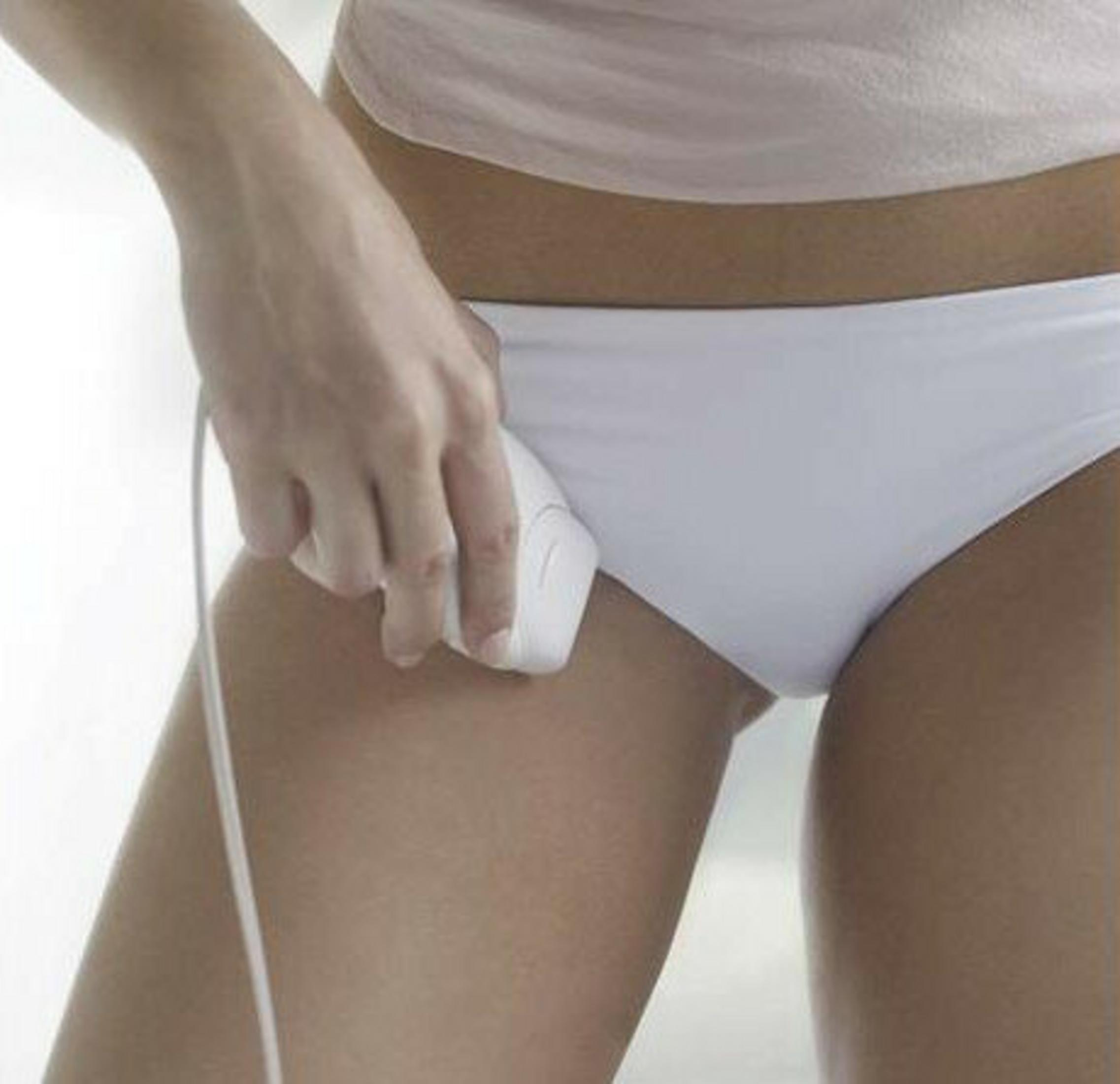 Osoba używa białego urządzenia do usuwania włosów na nodze. Ma na sobie białą bieliznę i jasnoróżową koszulę.