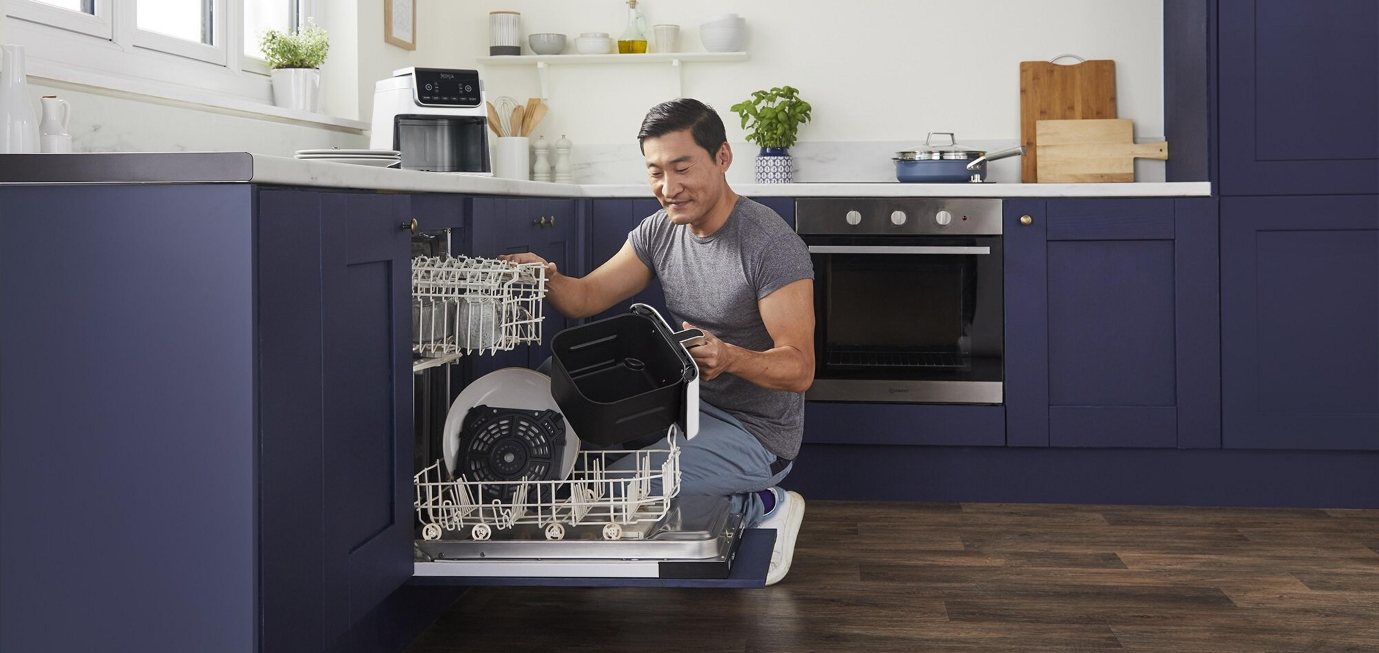 Mężczyzna w kuchni wkłada części frytownicy do zmywarki. Widoczne są niebieskie szafki i urządzenia.