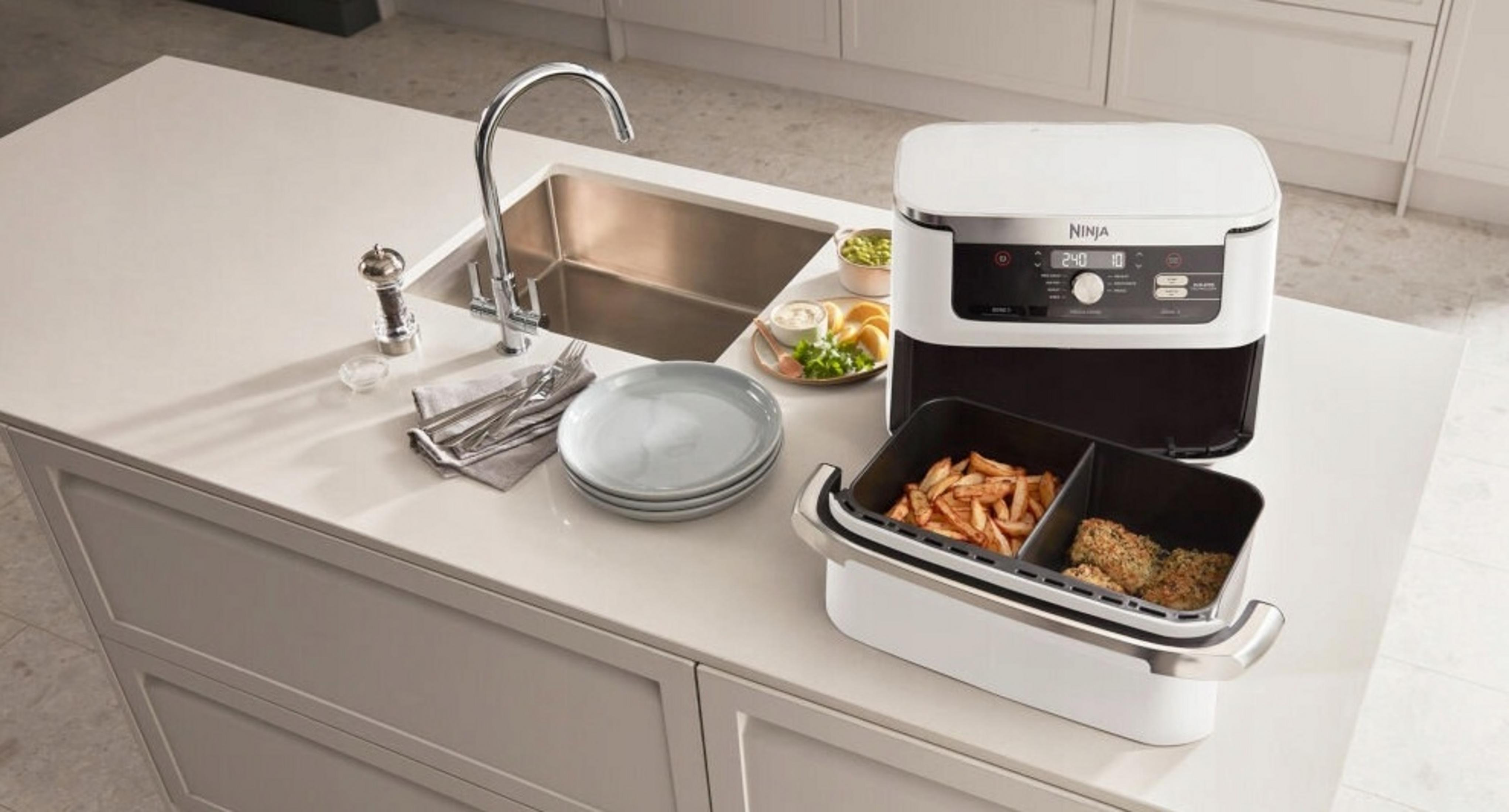 Blat kuchenny z frytownicą powietrzną Ninja, jedzeniem i naczyniami. Zlew w tle. Frytownica ma frytki i paluszki rybne.