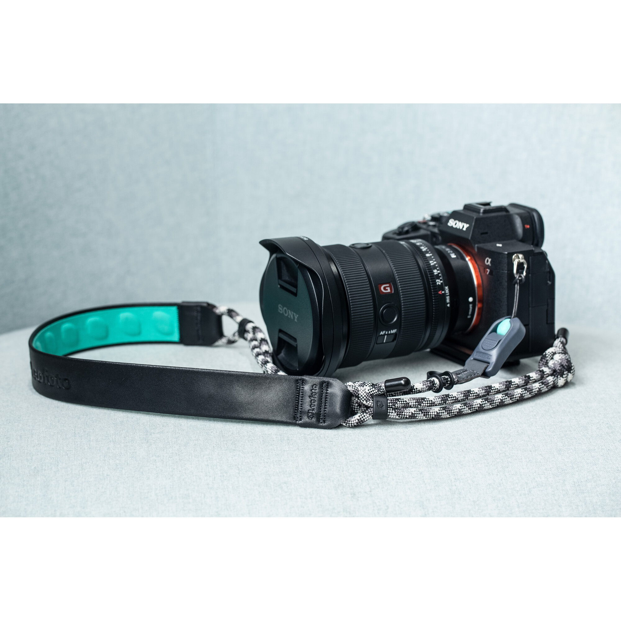 Eine schwarze Kamera mit Objektivdeckel liegt mit einem gemusterten Riemen. Der Riemen hat ein teal Kissen.