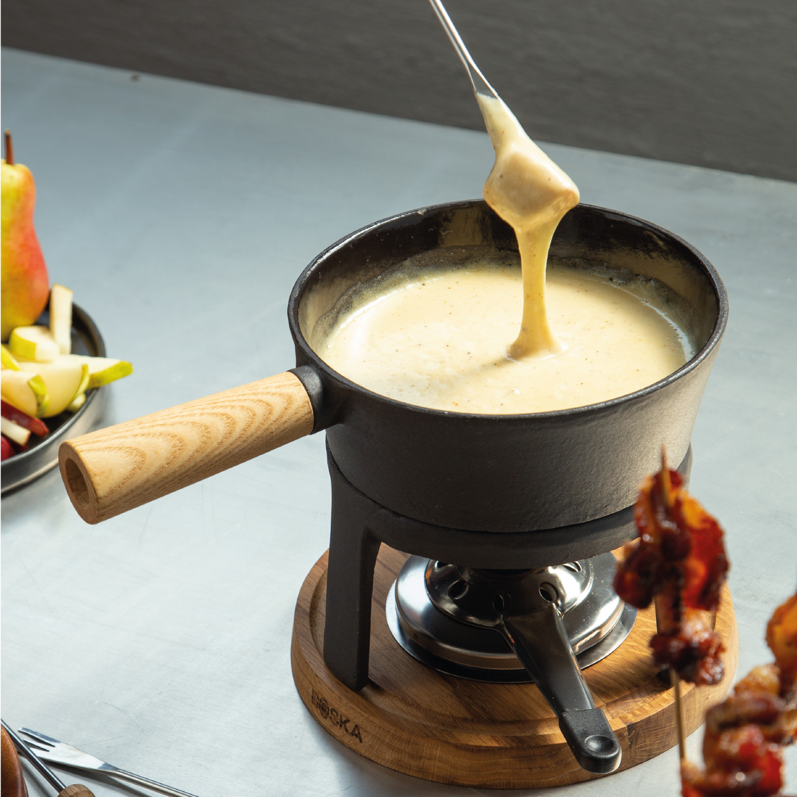 Garnek do fondue z wylanym serem. Jabłka, gruszki i szaszłyki mięsne z boku.