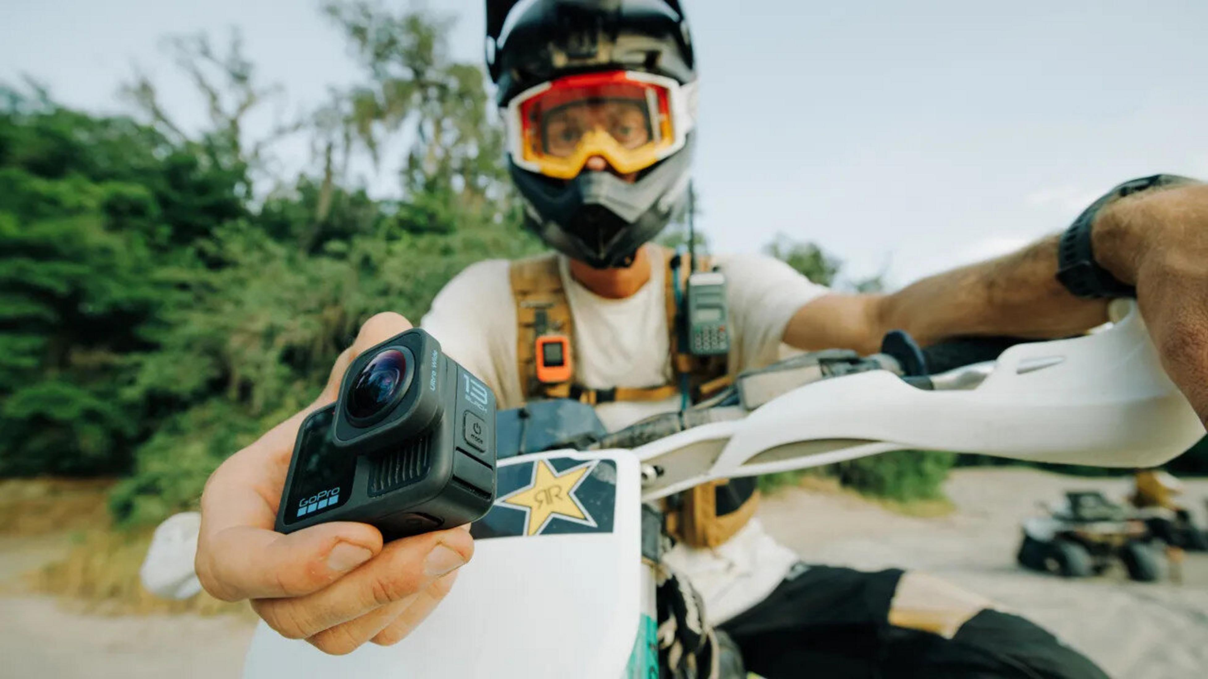Osoba w kasku trzyma kamerę GoPro na motocyklu. W tle drzewa i inny pojazd.