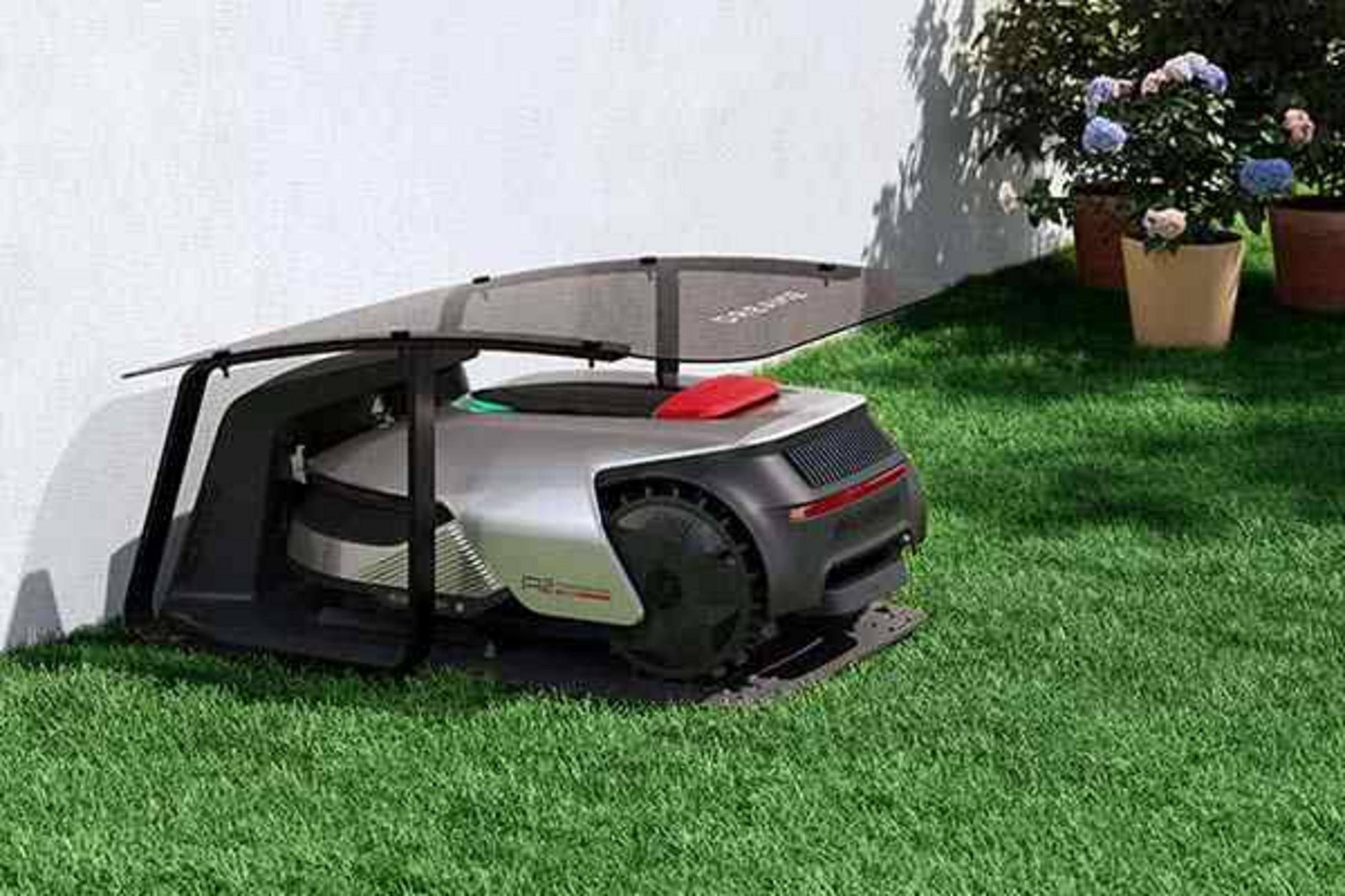Ein Roboter-Rasenmäher parkt unter einem Vordach auf grünem Gras. Blumen in Töpfen sind im Hintergrund sichtbar.