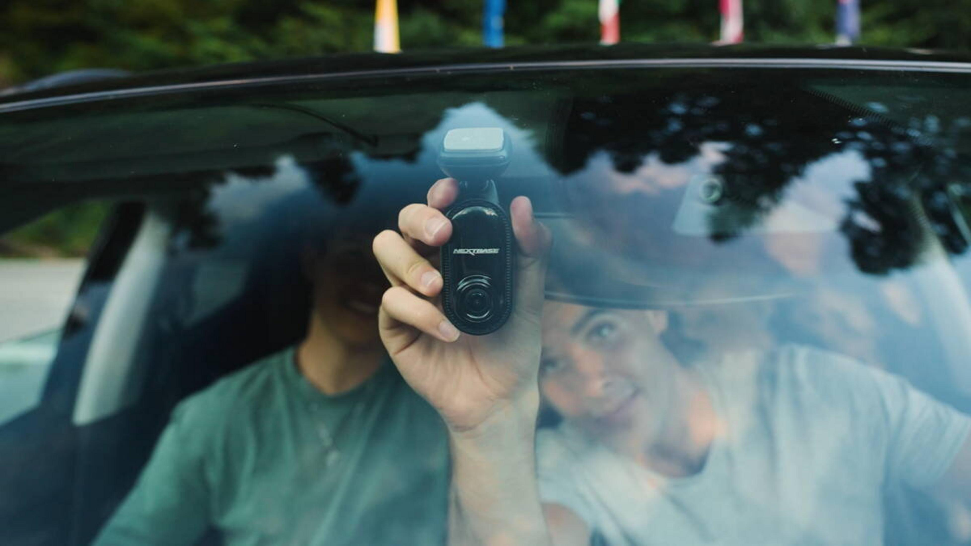 Eine Person hält eine schwarze Dashcam in einem Auto. Zwei Personen sind sichtbar. Grüne Bäume sind durch die Windschutzscheibe zu sehen.