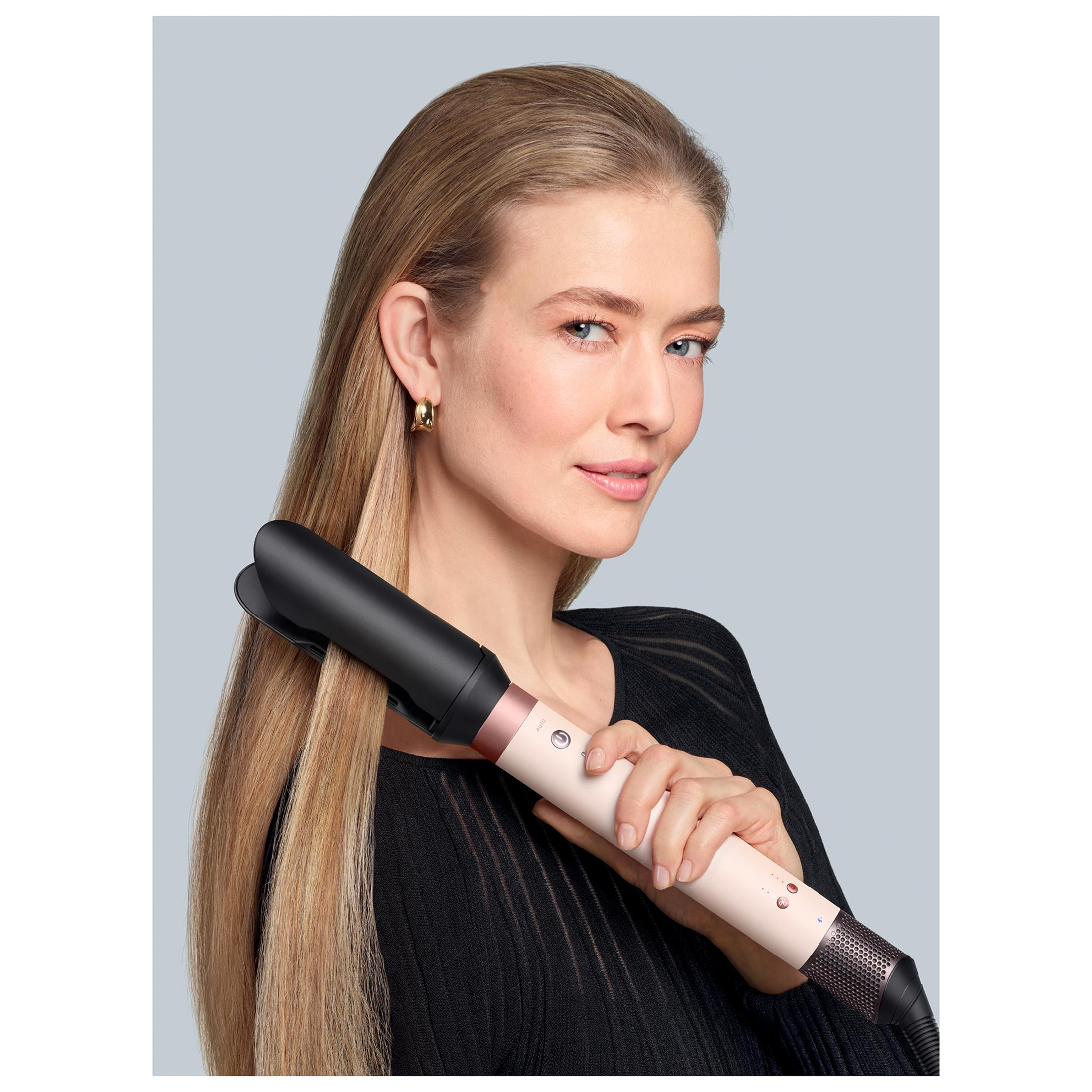 Kobieta z blond włosami trzymająca narzędzie do stylizacji włosów. Ma na sobie czarny top i złote kolczyki, uśmiecha się do kamery.