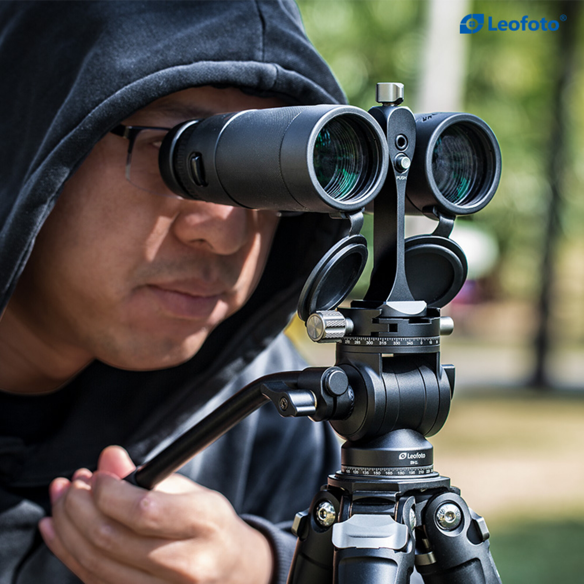 Eine Person blickt durch ein Fernglas, das auf einem Stativ montiert ist. Das Fernglas ist schwarz. Die Person trägt eine Brille und eine Kapuze.
