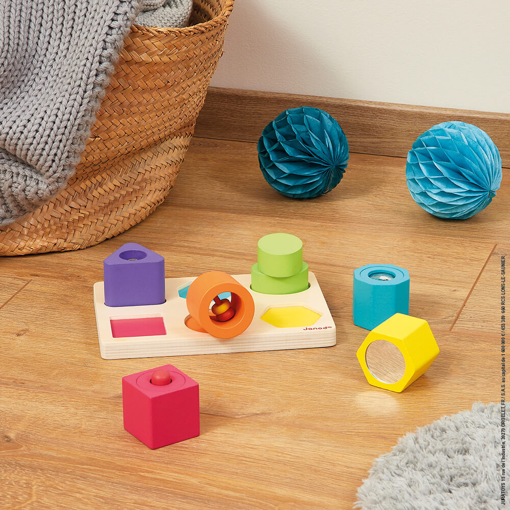 Clasificador de formas de madera con bloques coloridos, dos bolas de papel azules y una cesta con una manta en un suelo de madera.