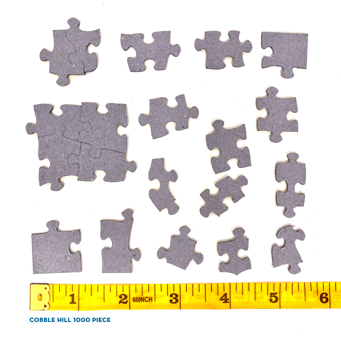 Verstreute graue Puzzleteile auf Weiß. Ein gelbes Maßband mit Zollmarkierungen und dem Text 'Cobble Hill 1000 Piece' ist vorhanden.