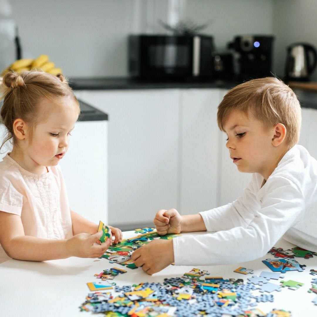 Twee kinderen leggen een puzzel aan een tafel. De een houdt een stuk vast, de ander reikt naar een ander stuk. Keukenachtergrond.