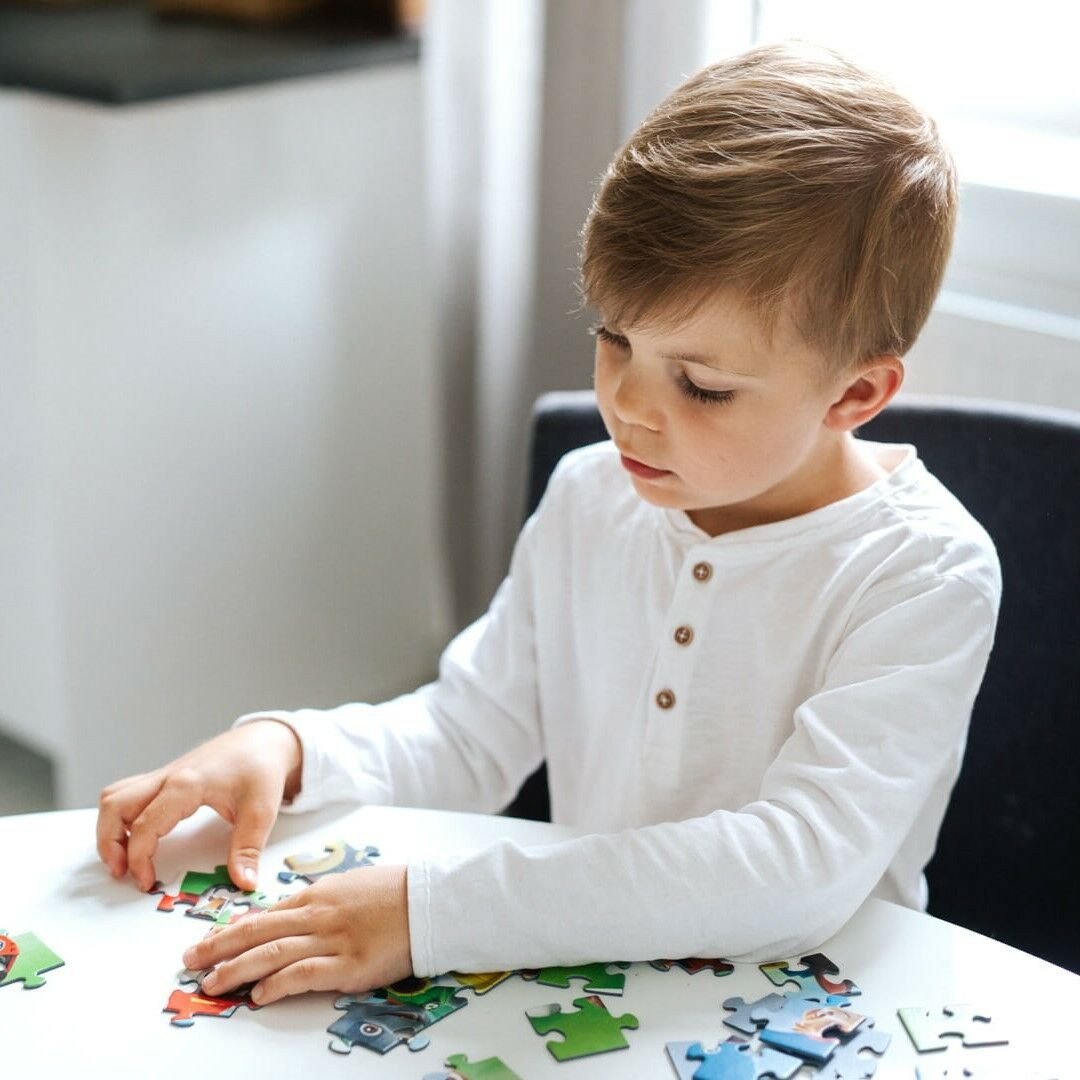 Een jongen met blond haar zit aan een witte tafel en legt een puzzel. Hij draagt een wit shirt.