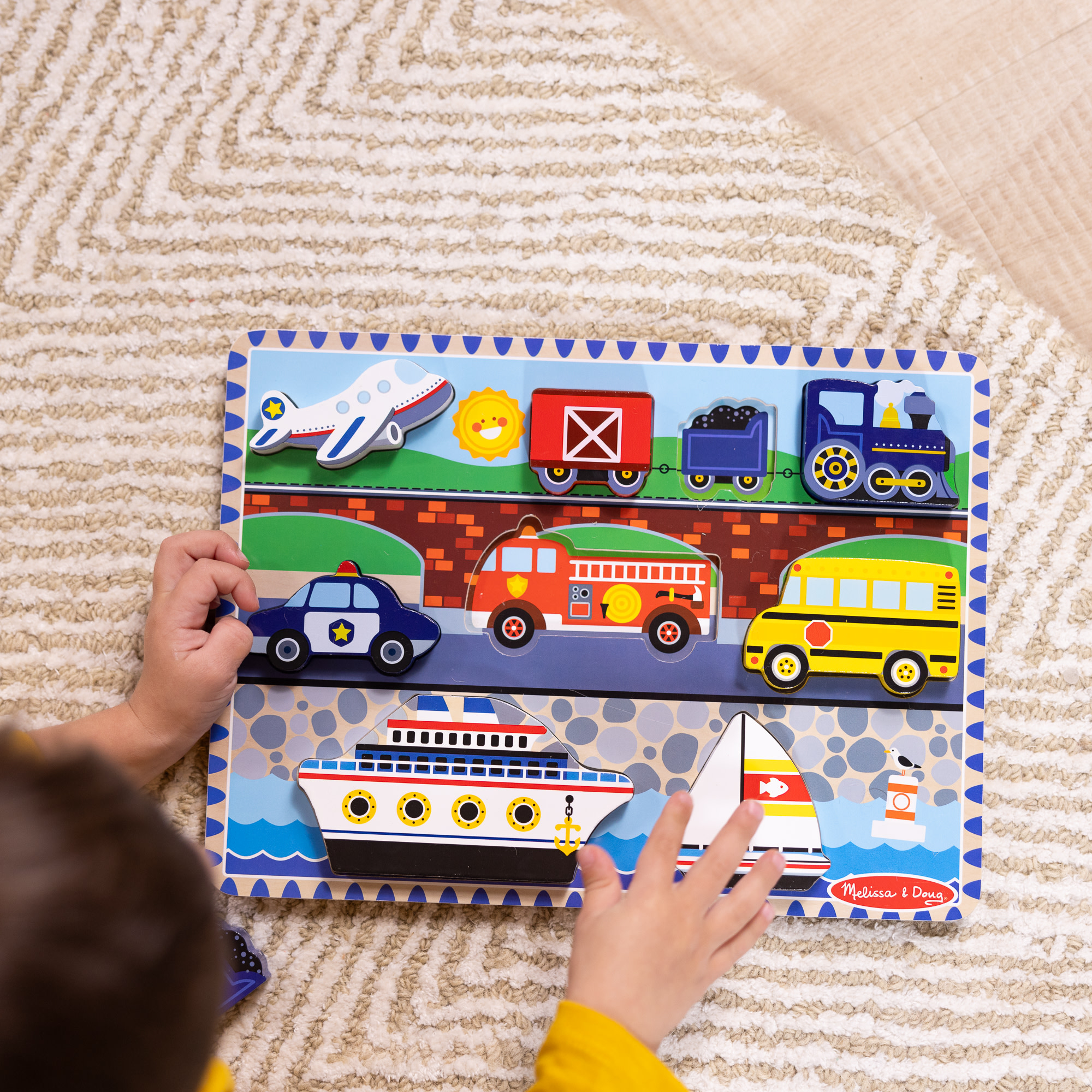 Ein Kind spielt mit einem Holzpuzzle mit Fahrzeugen: Flugzeug, Polizeiauto, Feuerwehrwagen und Segelboot.