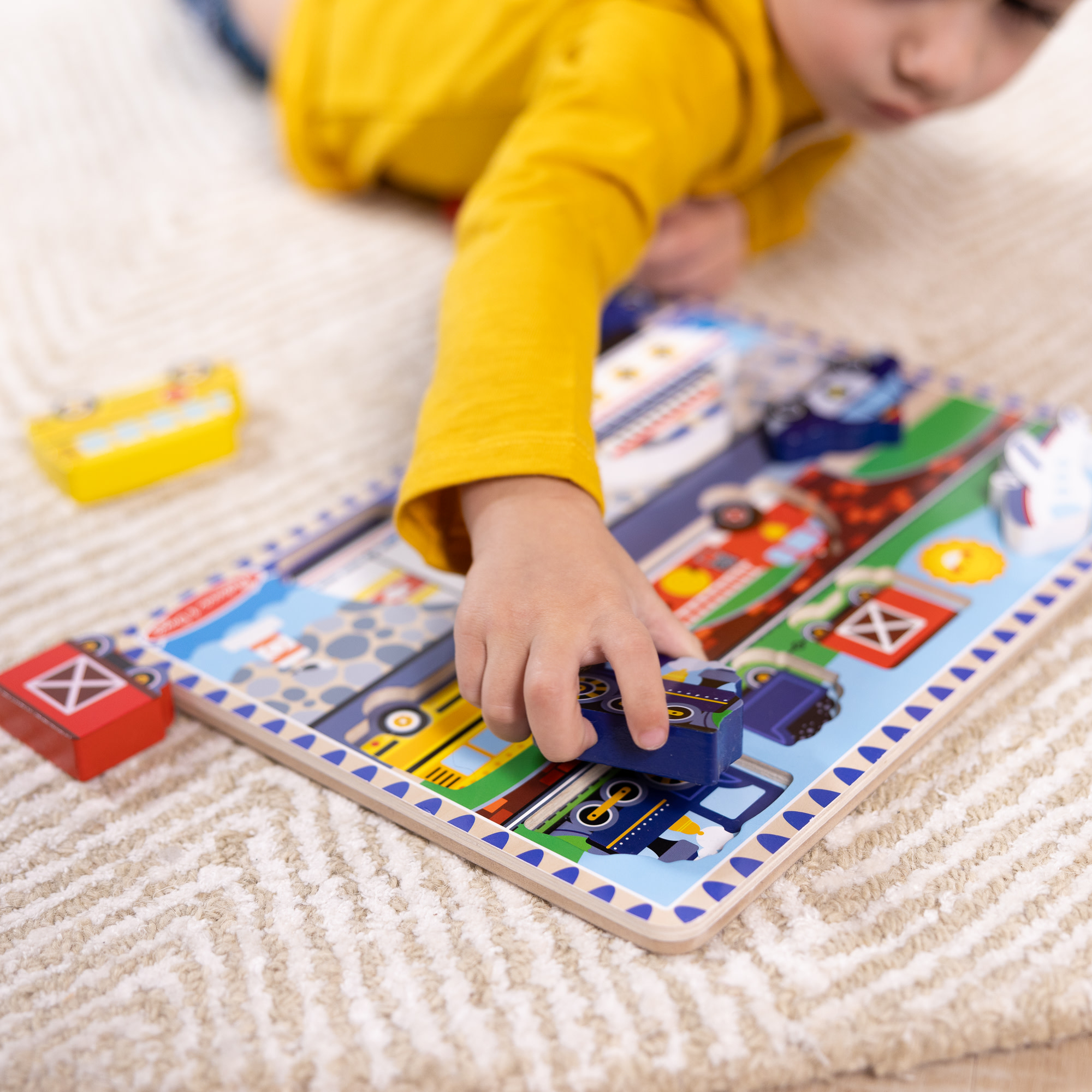 Ein Kind spielt mit einem Holzpuzzle von Fahrzeugen. Die Hand des Kindes platziert einen blauen Lastwagen. Gelbes Hemd.