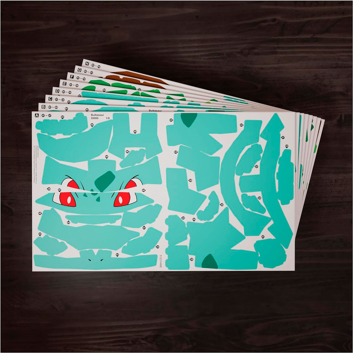 Blätter mit Bulbasaur-Ausschnitten auf dunkler Holzoberfläche. Die Blätter zeigen Bulbasaur's Gesicht und Körperteile in verschiedenen Grüntönen.