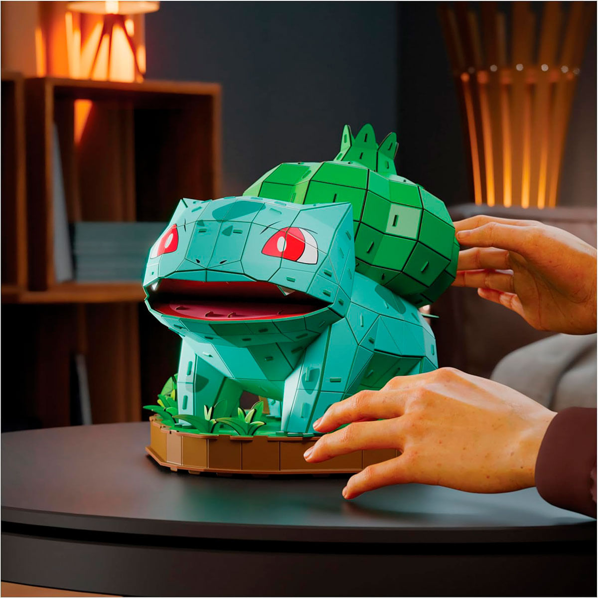 Ein türkisfarbenes Bulbasaur-Modell steht auf einem Tisch. Hände sind in der Nähe des Modells. Der Hintergrund hat eine dunkle Wand und Holzregale.