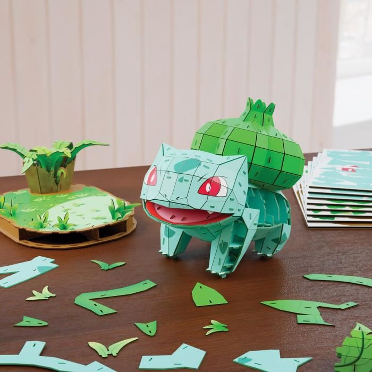 Ein zusammengebautes und auseinandergebautes Bulbasaur-Papierhandwerk und eine Pflanze auf einer Plattform stehen auf einem Holztisch. Ein Stapel Karten ist im Hintergrund.
