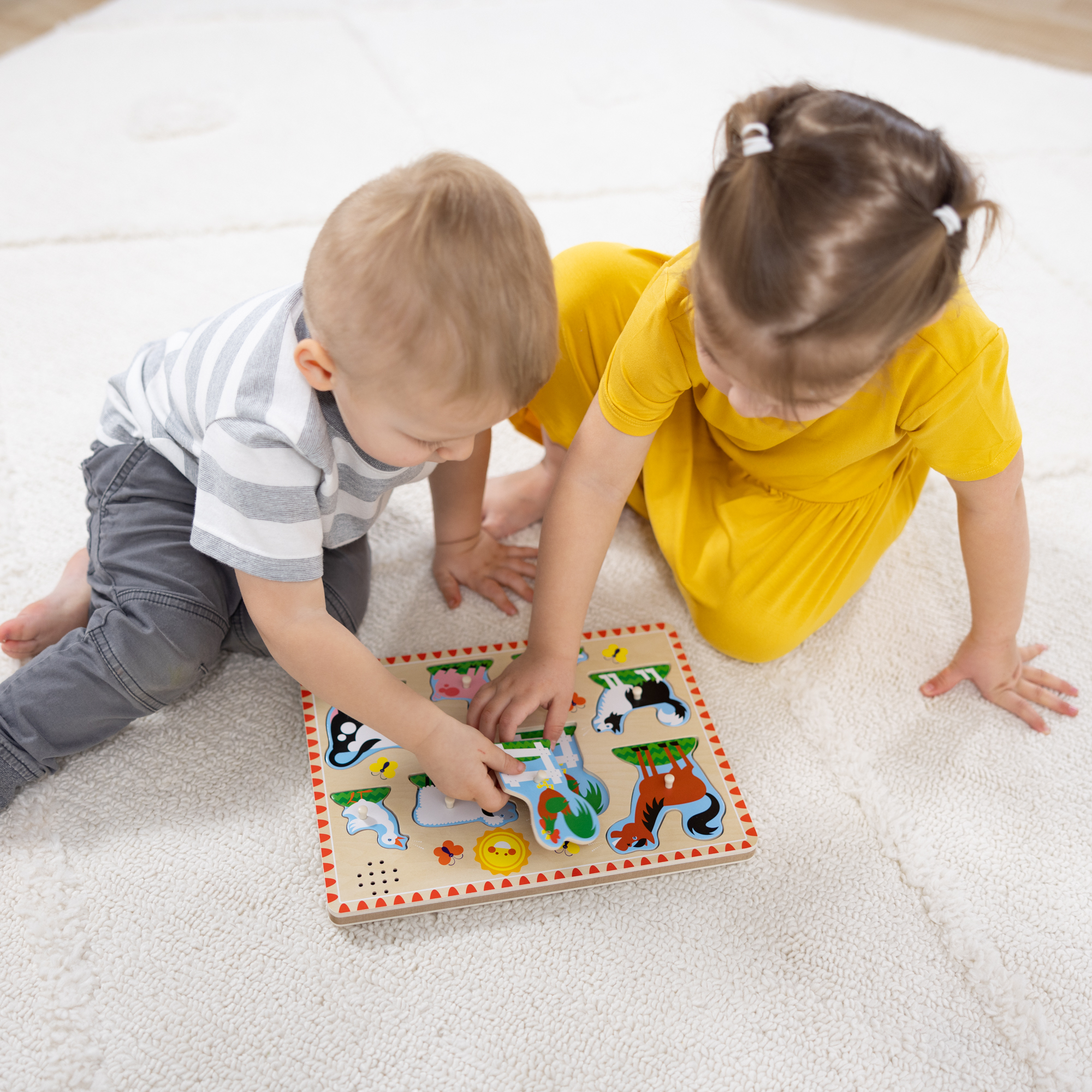 Zwei Kinder spielen auf einem weißen Teppich mit einem Holzpuzzle. Eines trägt ein gestreiftes Hemd, das andere ein gelbes Kleid.