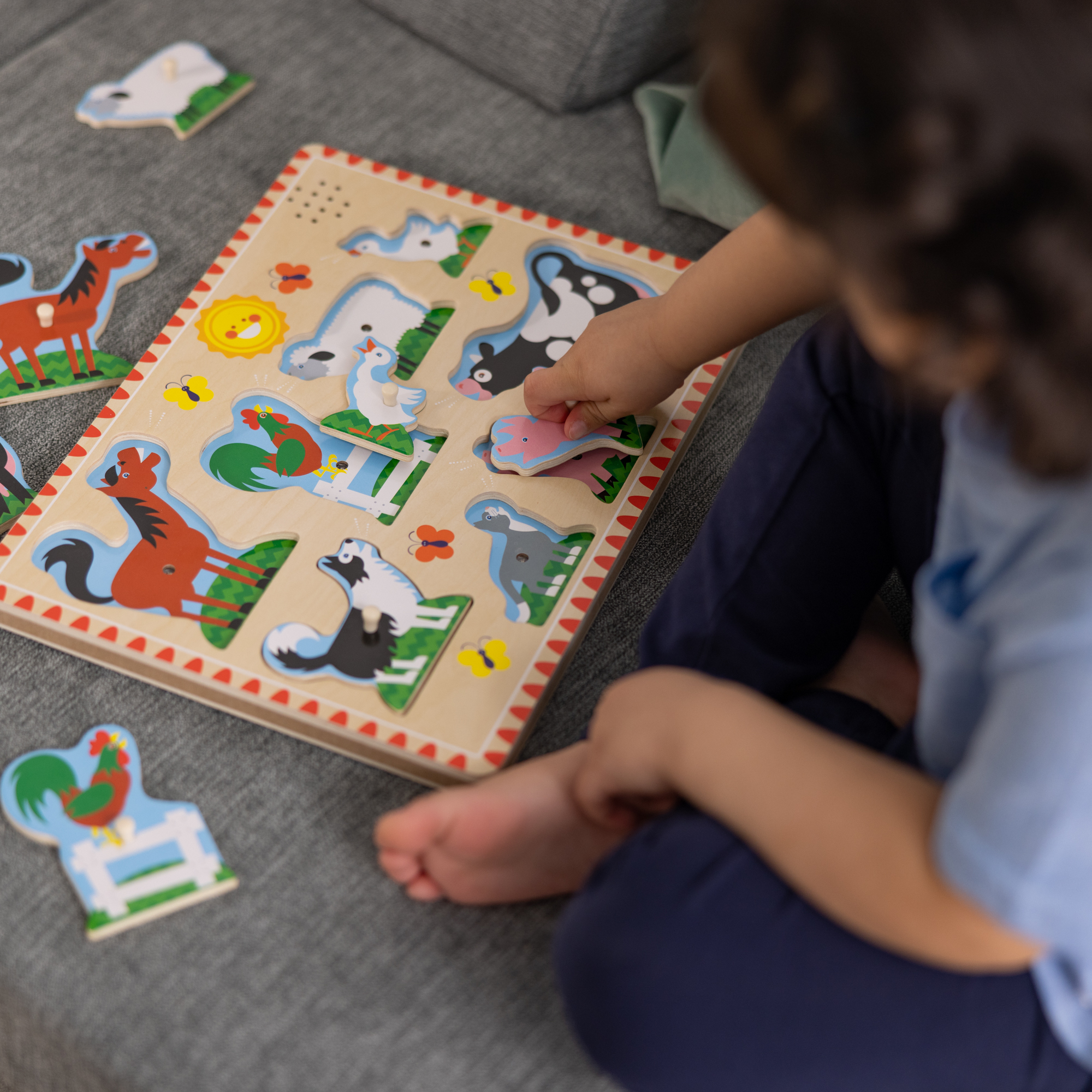 Ein Kind spielt mit einem Holzpuzzle mit Bauernhoftieren auf einer grauen Oberfläche. Das Kind legt ein Schwein-Teil ein.