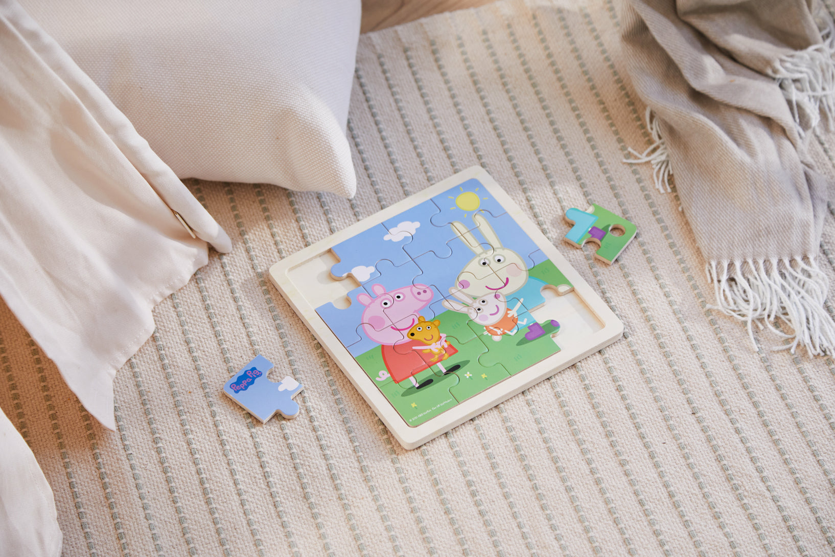 Ein Peppa Wutz Puzzle auf einem Teppich. Das Puzzle ist teilweise zusammengesetzt, zwei Teile liegen daneben. Ein Kissen und eine Decke sind auch vorhanden.