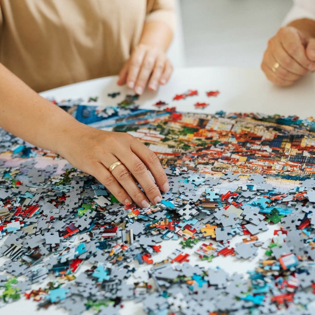 Mains avec des bagues assemblant un puzzle coloré sur une table blanche. Des pièces éparpillées entourent l'image partiellement complétée.