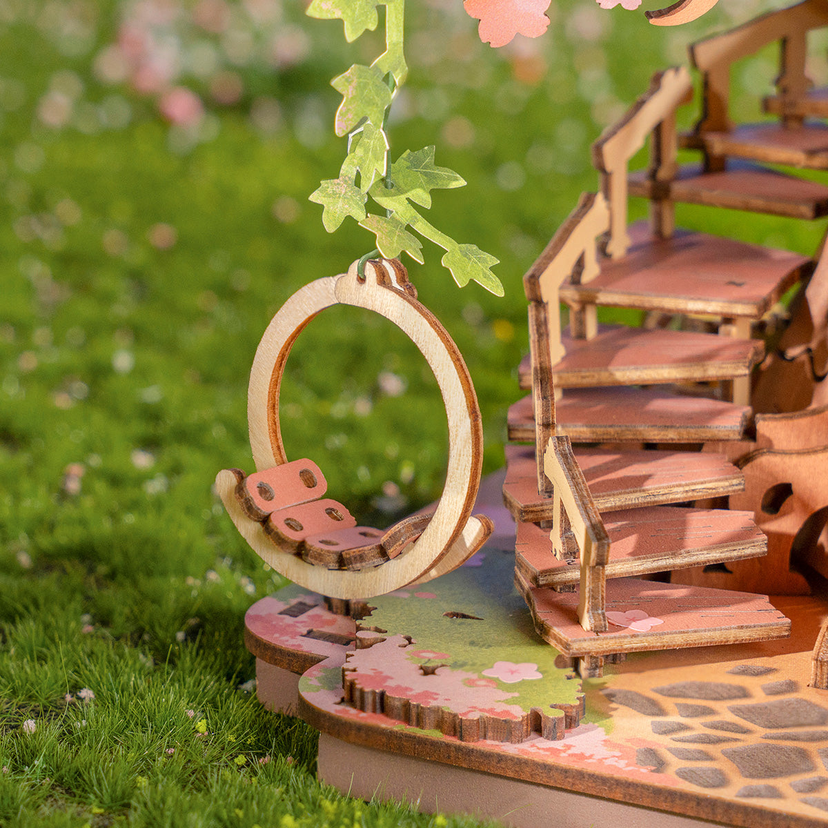 Miniaturgarten mit Holzschaukel und Treppe, mit grünem Gras und rosa Blumen im Hintergrund.