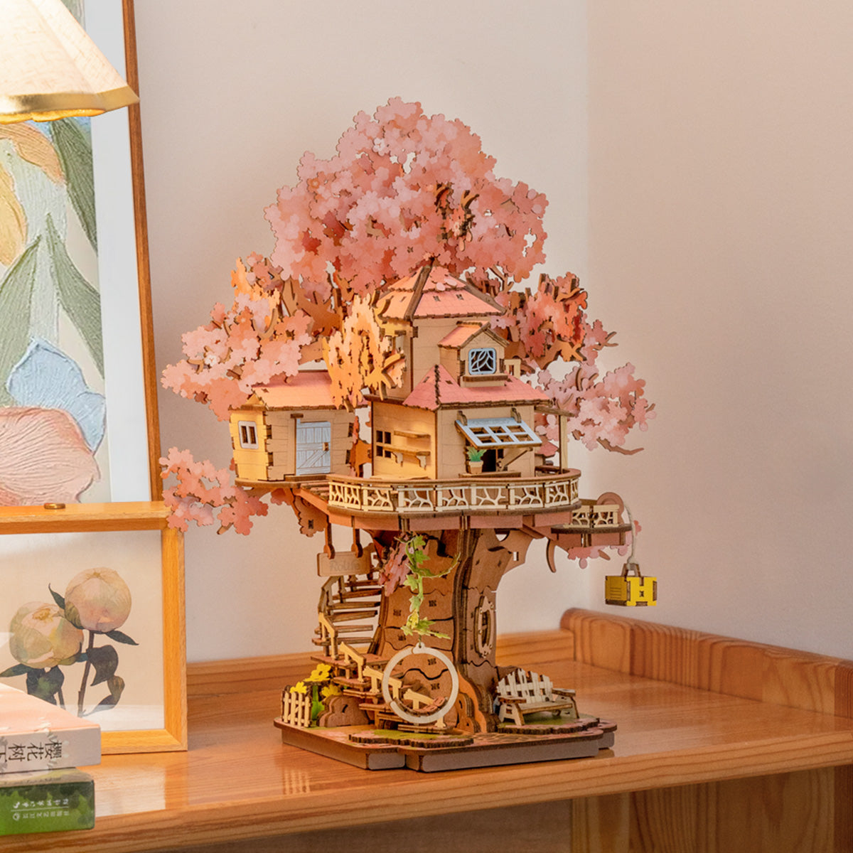 Ein hölzernes Baumhausmodell mit rosa blühenden Ästen, auf einem Holztisch. Andere Objekte sind eine Lampe und ein Bilderrahmen.