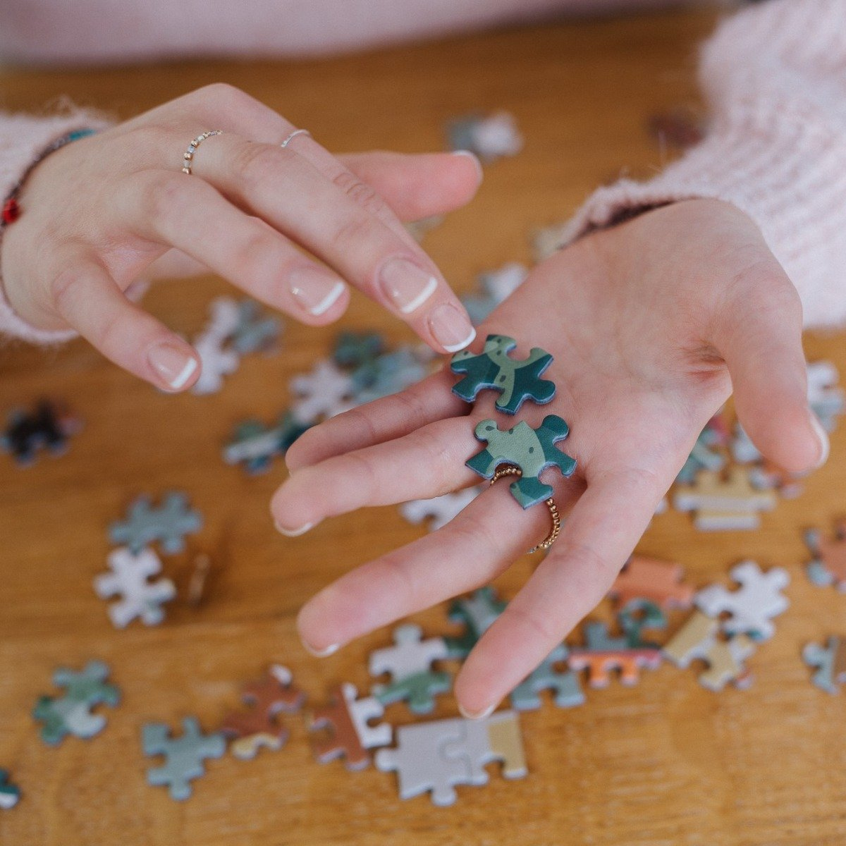 Hände halten zwei Puzzleteile. Andere Teile liegen auf einem Holztisch verstreut. Die Person trägt einen rosa Pullover und Ringe.
