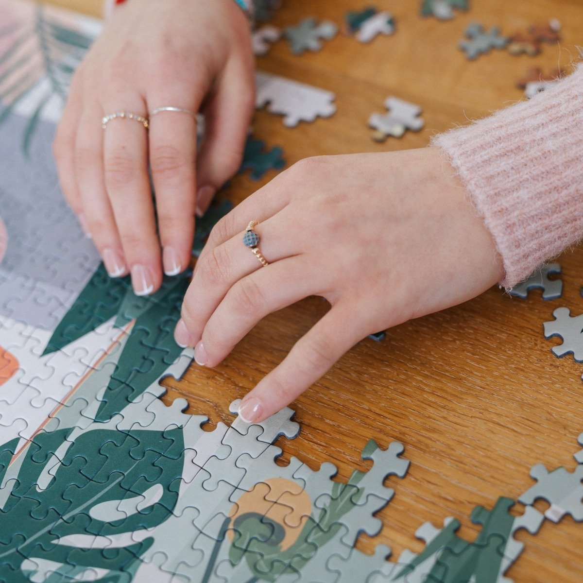 Hände mit Ringen setzen ein Puzzle zusammen. Das Puzzle liegt auf einem Holztisch. Einige Teile sind bereits platziert, andere verstreut.