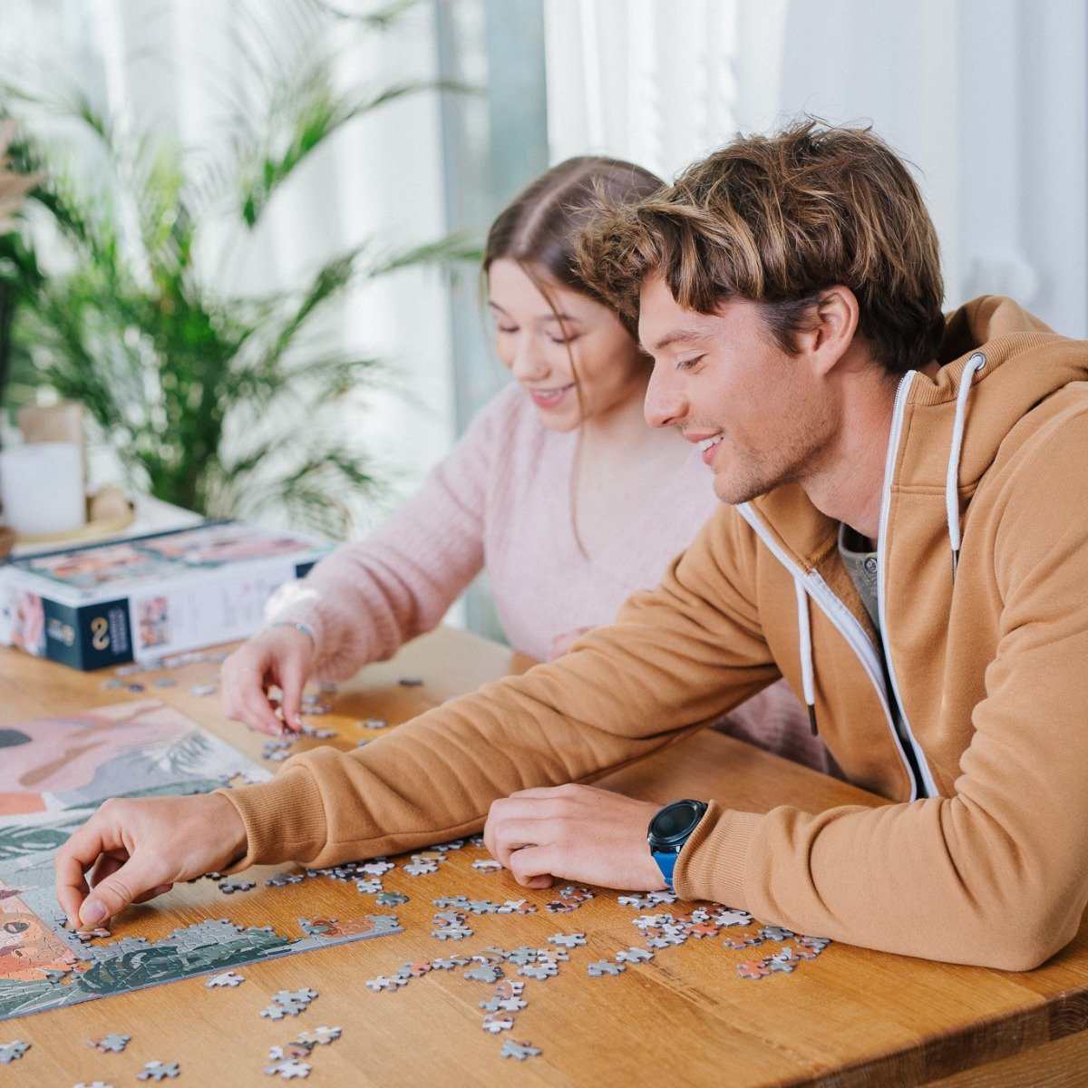 Ein Mann und eine Frau puzzeln an einem Tisch. Der Mann trägt einen braunen Kapuzenpullover. Eine Schachtel und eine Pflanze sind im Hintergrund.