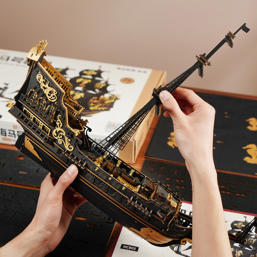 Une personne tient une maquette de bateau en bois noir et or. Le navire est détaillé avec des mâts, des voiles et des éléments décoratifs.