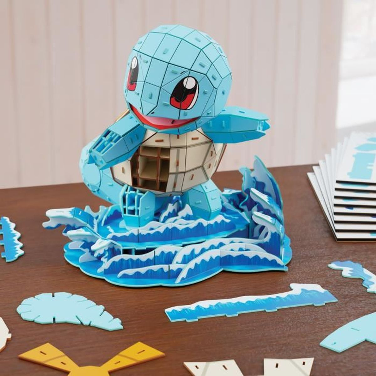 Ein blaues Squirtle-3D-Puzzle auf einer Wellenbasis. Verstreute Puzzleteile liegen auf einem Holztisch. Der Hintergrund ist eine helle Wand.