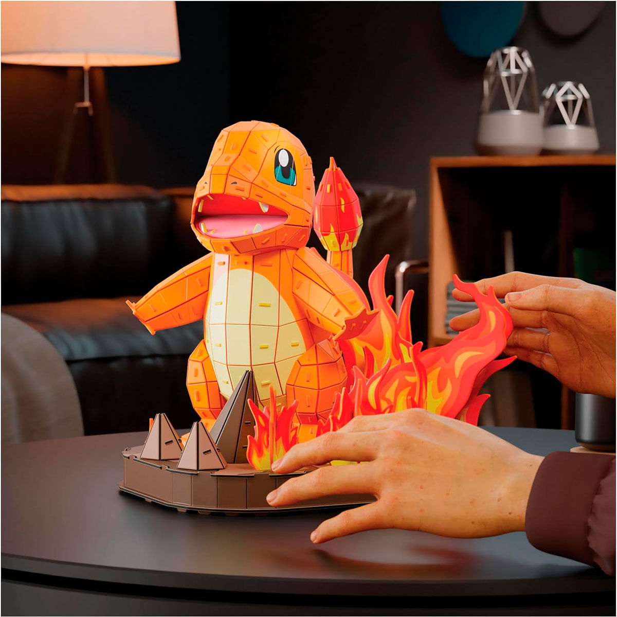 Un modello di Charmander con fiamme. Le mani posizionano i pezzi della fiamma su un tavolo. Lo sfondo include una lampada e degli altoparlanti.