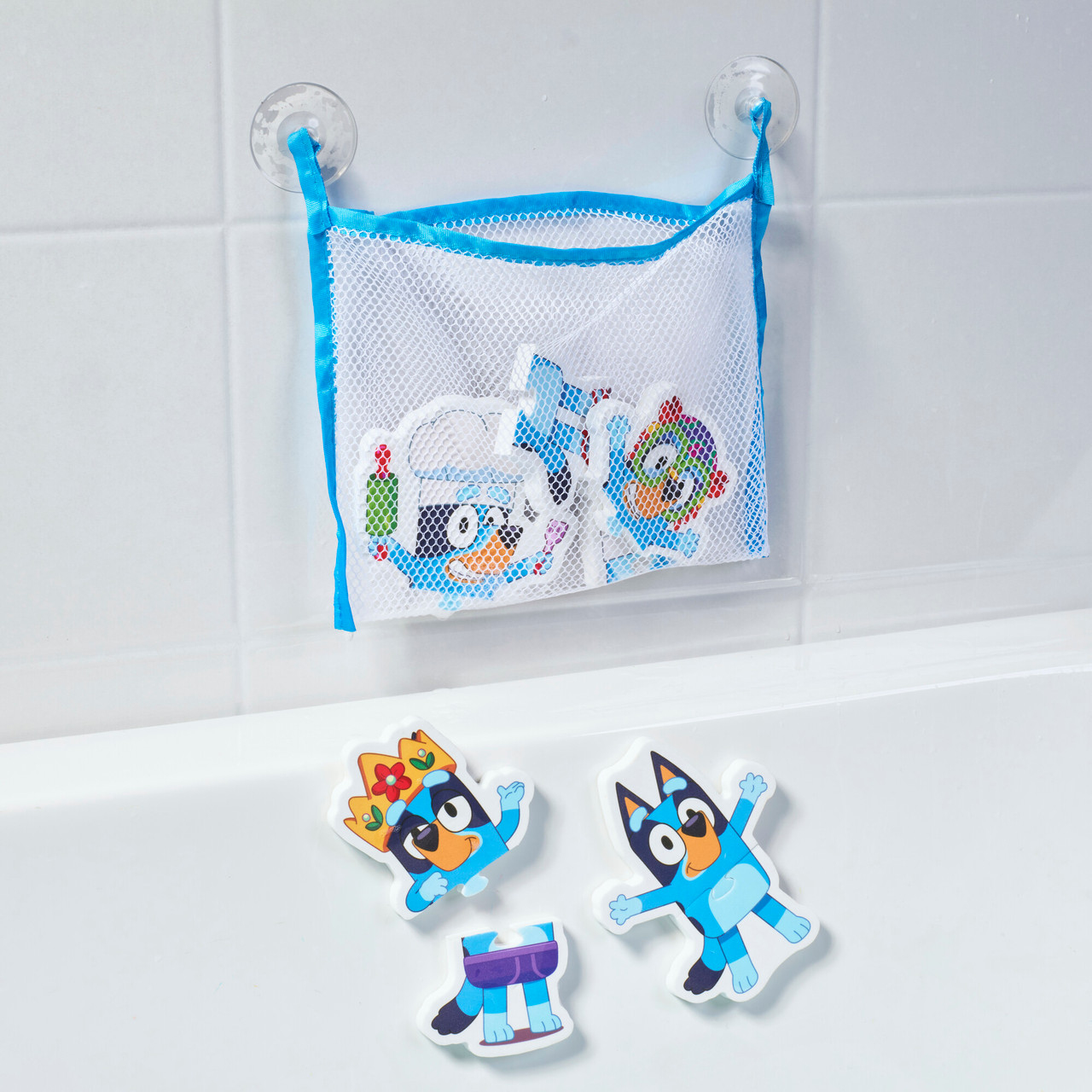 Eine Netztasche mit Bluey-Badespielzeug hängt an einer gefliesten Wand. Drei Spielzeuge liegen am Wannenrand. Blau, weiß und orange.
