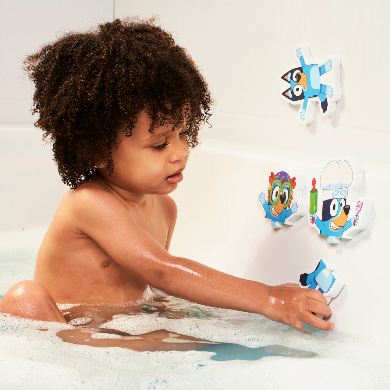 Ein Kind in einer Badewanne greift nach einem Bluey-Charakter-Badespielzeug. Andere Spielzeuge sind an der Wanne befestigt.