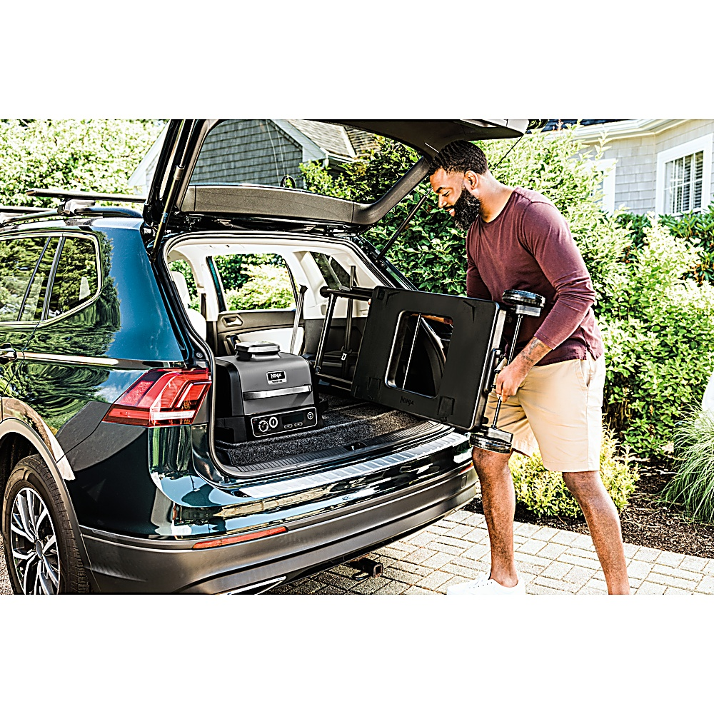 Mężczyzna ładuje czarnego grilla i akcesoria do bagażnika samochodu. Samochód jest ciemnozielony, zaparkowany blisko domu z zielenią.