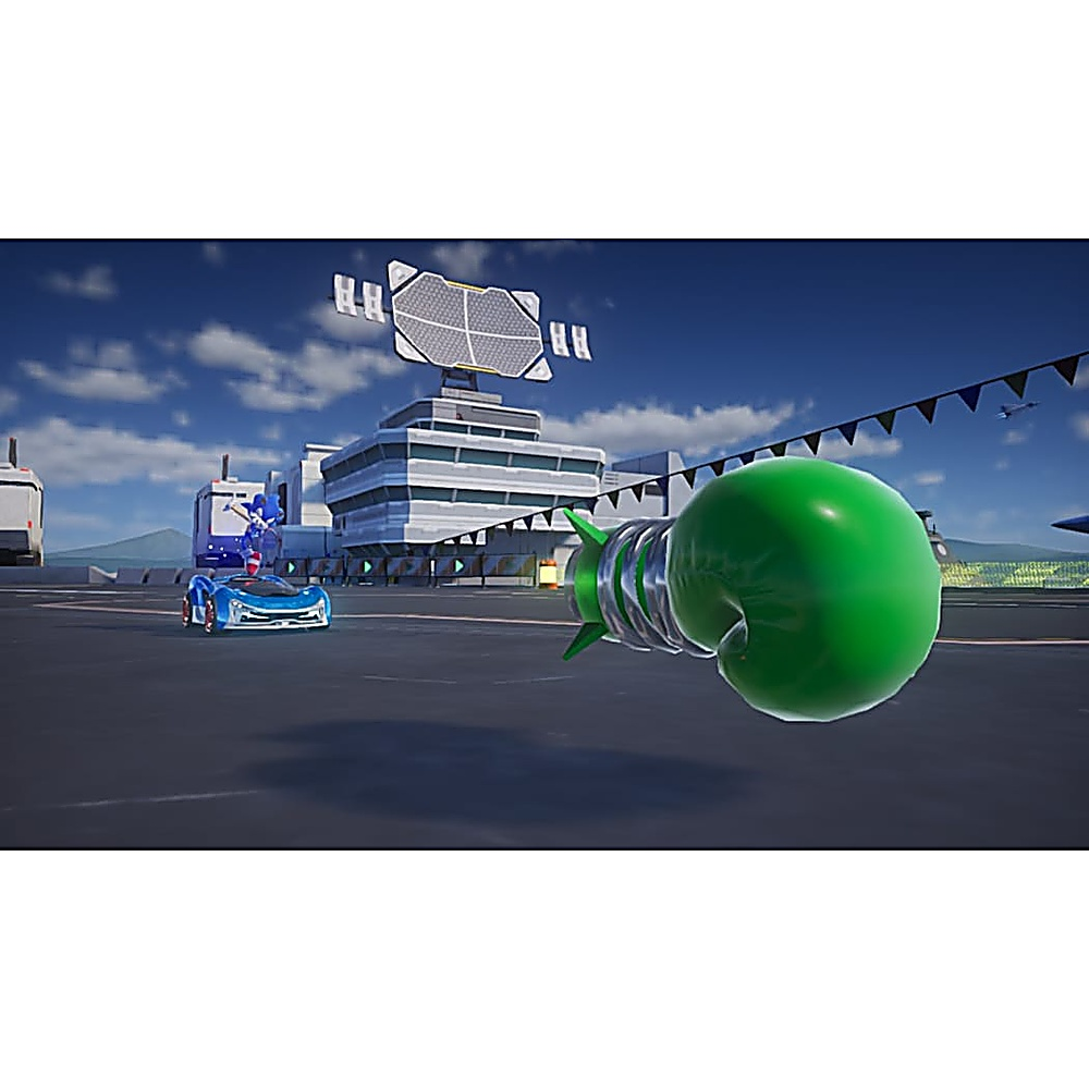 Sonic w niebieskim samochodzie, zielona rękawica bokserska po prawej, budynek z radarem, błękitne niebo z chmurami, flagi.