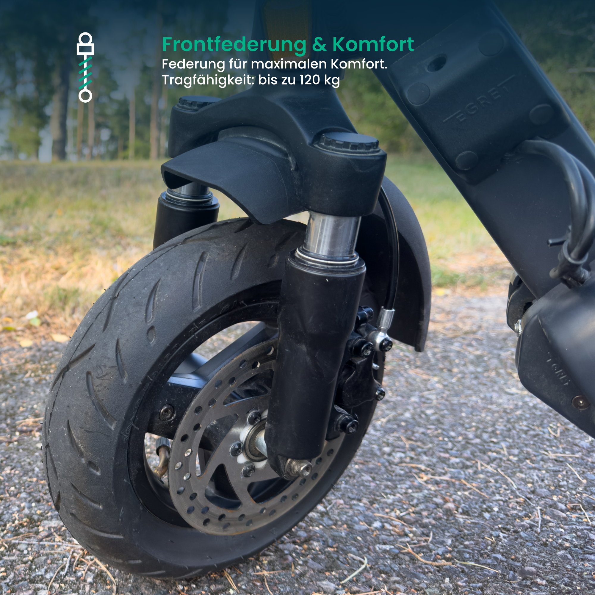 Nahaufnahme eines schwarzen Rollerreifens mit Federung. Der Hintergrund ist ein verschwommener Wald. Der Roller steht auf einem Kiesweg.