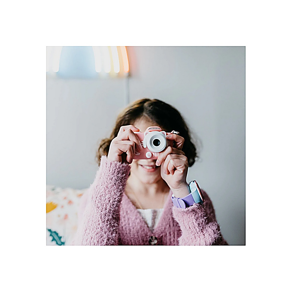 Una niña sonríe mientras sostiene una cámara rosa. Lleva un suéter rosa y un reloj. Una pared con luz está en el fondo.