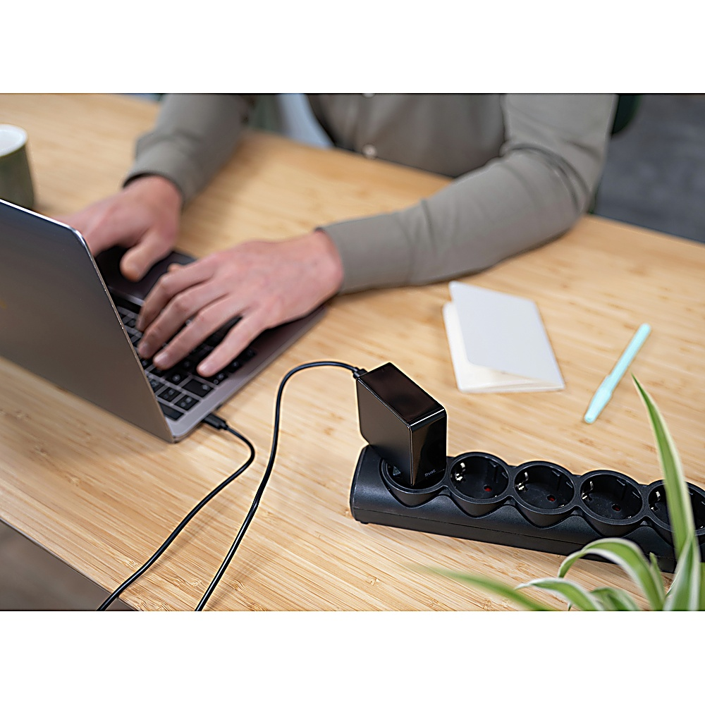 Una persona usa un laptop. Una ciabatta e un caricabatterie neri sono sulla scrivania di legno. Sono visibili anche una penna e un taccuino.