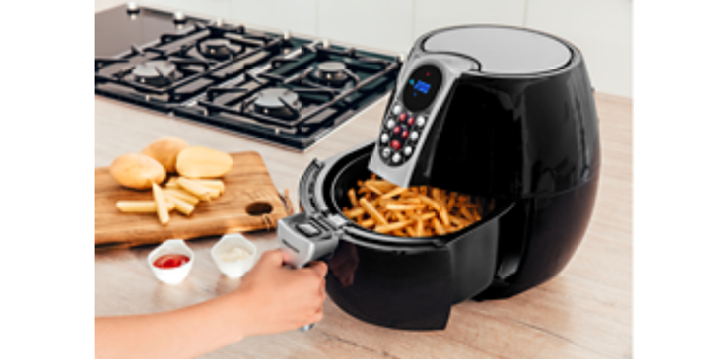 Ein Airfryer mit Pommes Frites, Kartoffeln auf einem Schneidebrett und kleine Schalen mit Soße sind sichtbar.