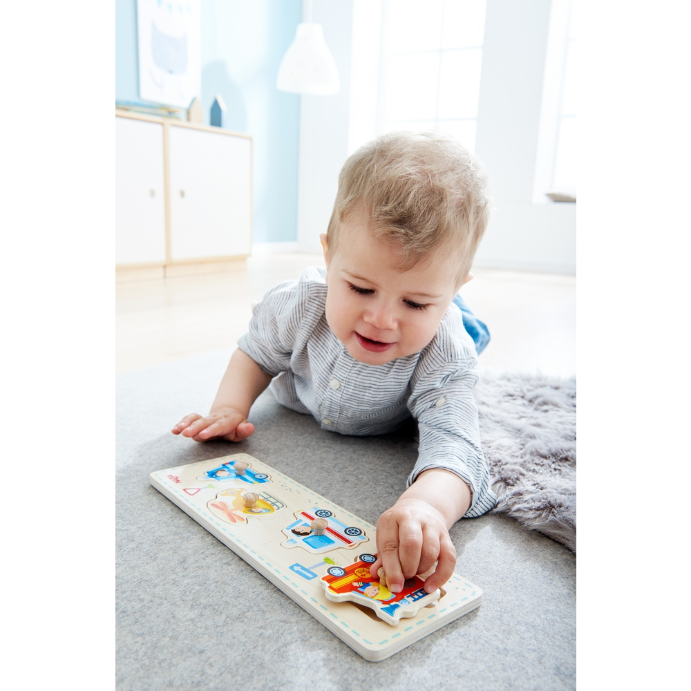 Ein Baby spielt auf einem grauen Teppich mit einem Holzpuzzle. Das Puzzle zeigt Fahrzeugformen. Im Hintergrund befindet sich ein grauer Teppich.