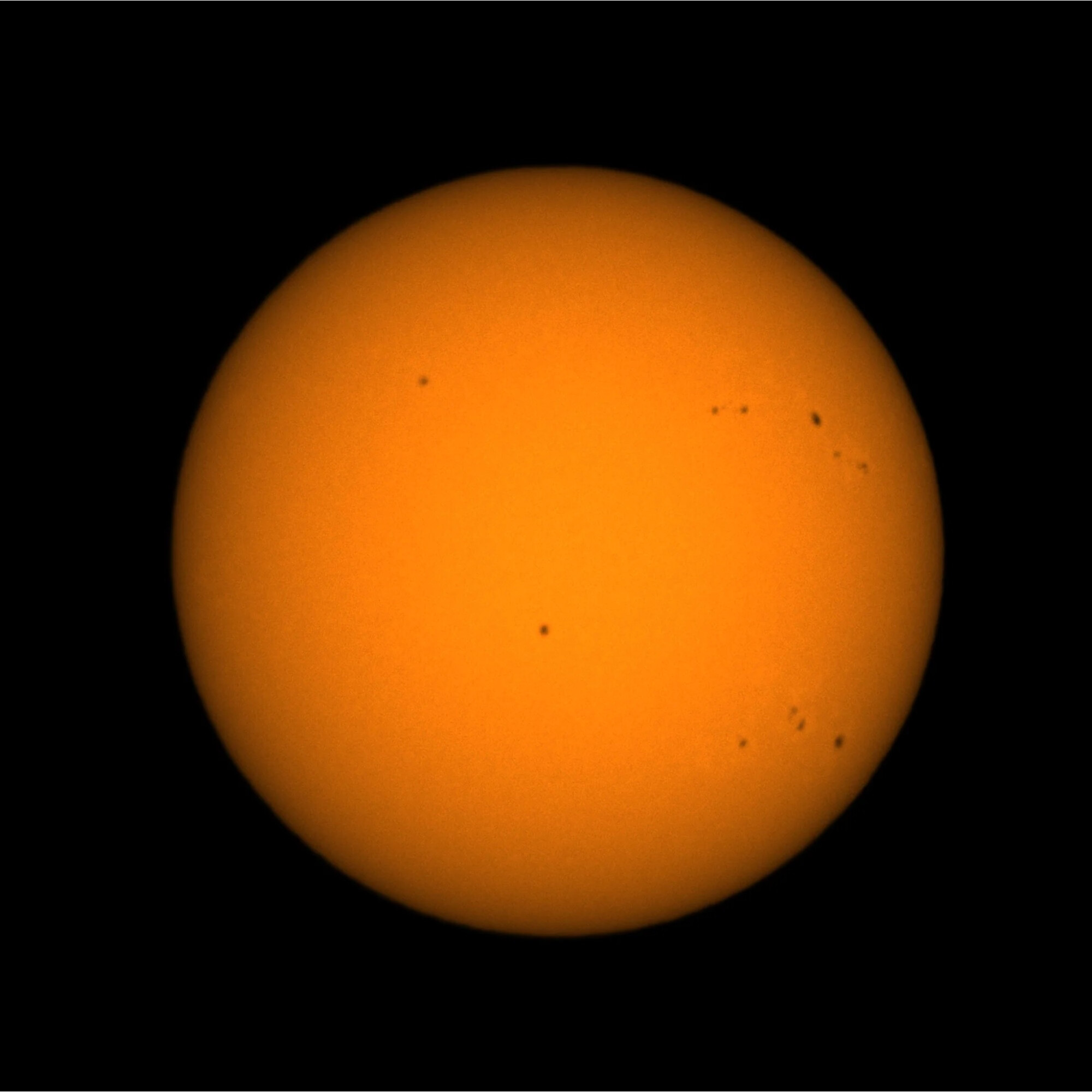 Eine leuchtend orangefarbene Kugel mit dunklen Flecken auf schwarzem Hintergrund, ähnelt der Sonne.