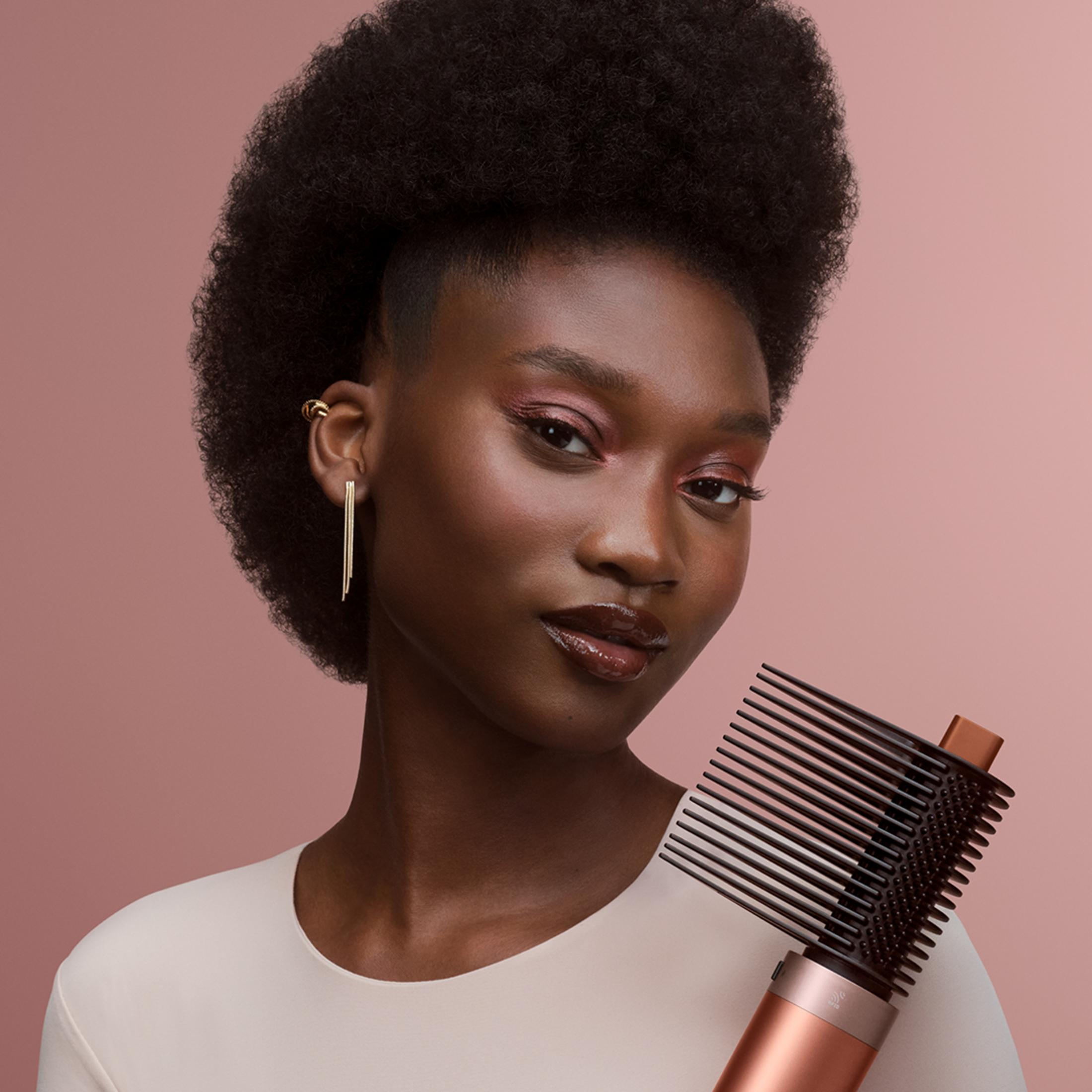 Una donna afro tiene in mano un arricciacapelli. Indossa trucco e orecchini d'oro, sfondo rosa.